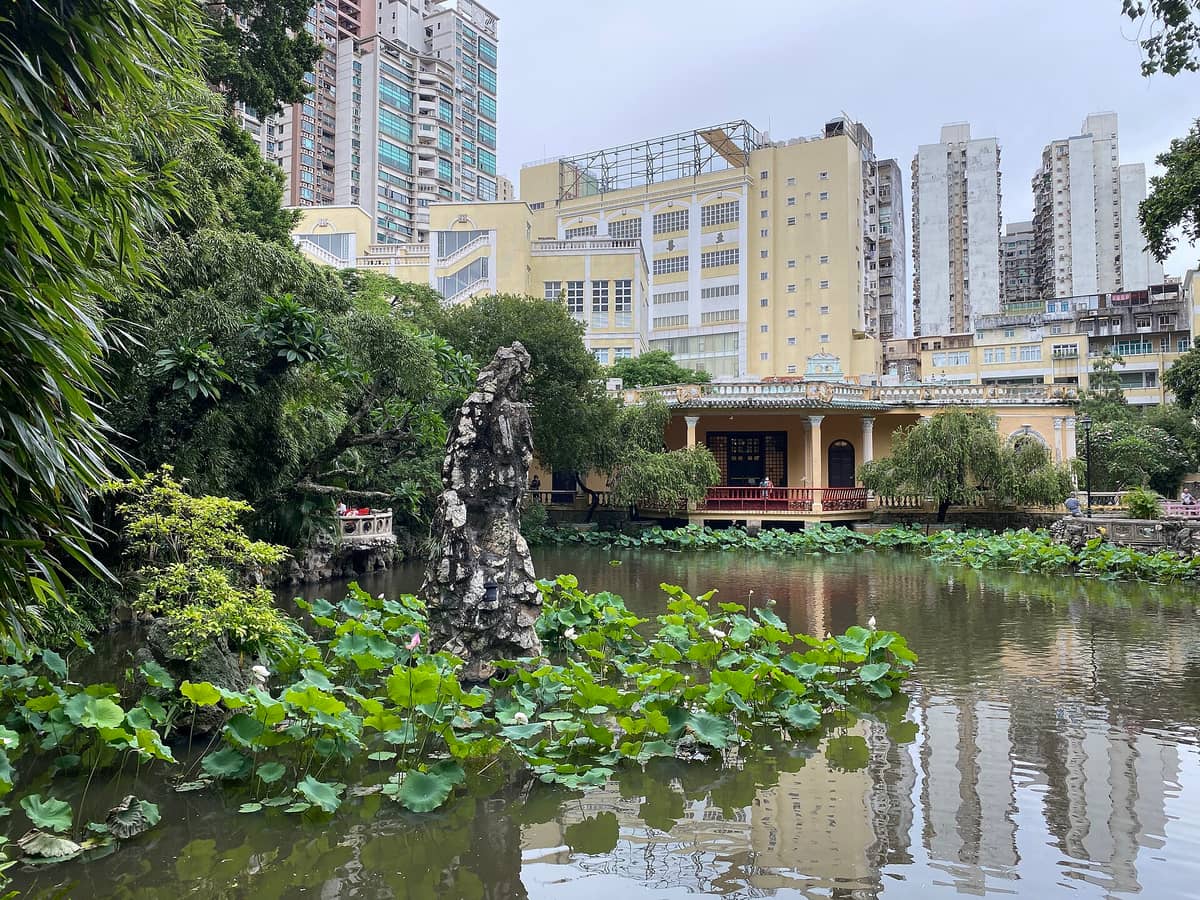 Der Lou Lim Ieoc Garden in Macau mit üppigem Lotusteich, klassischem chinesischem Pavillon und gelbem Herrenhaus im Hintergrund, dahinter die Hochhäuser der Stadt.