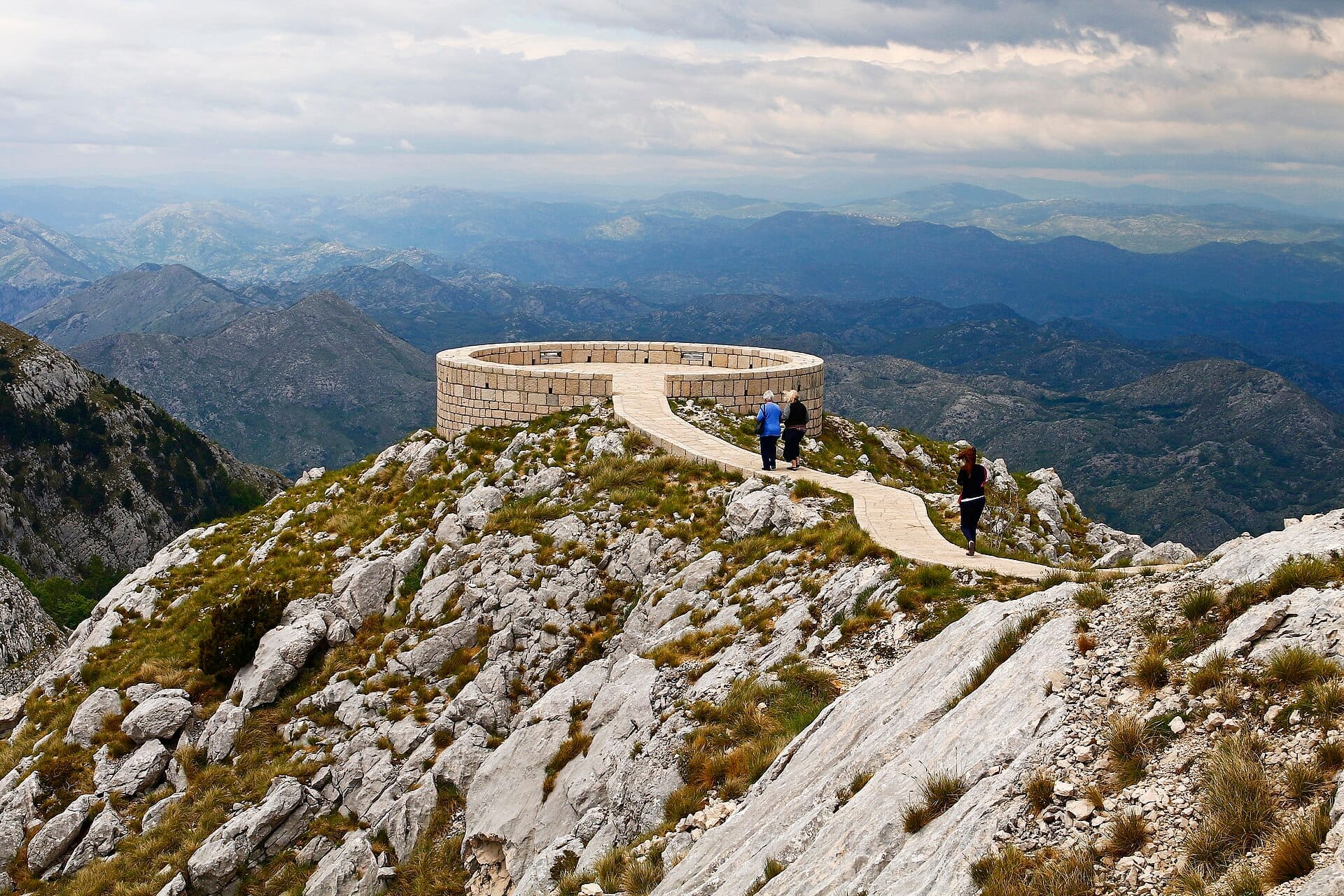 Belvedere circolare in pietra sulla cima del monte Lovćen, circondato da spettacolari vette rocciose e cieli nuvolosi, con tre visitatori che si avvicinano lungo un sentiero stretto.