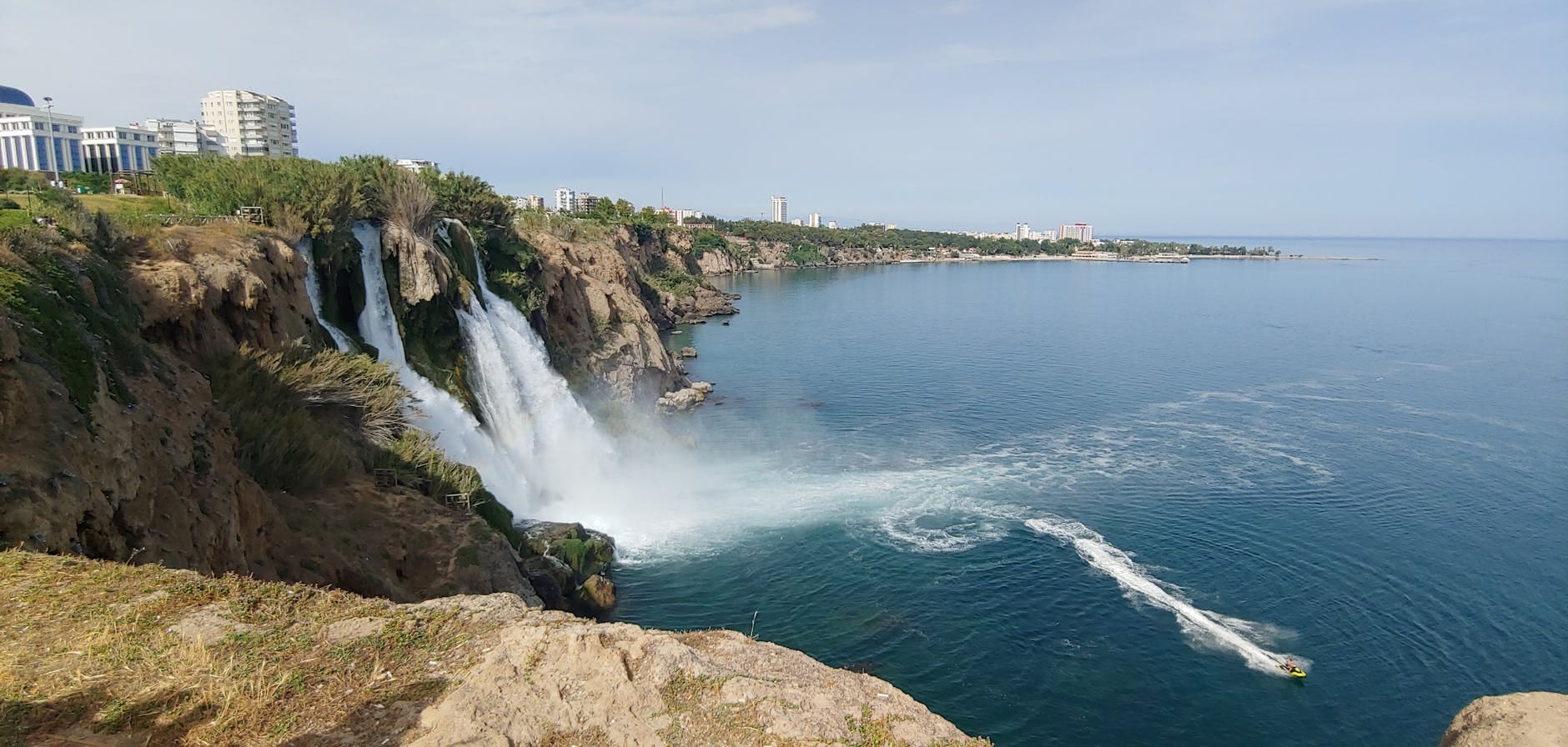 Vista panoramica delle Cascate Inferiori del Düden che scendono da una falesia nel Mar Mediterraneo, con edifici sullo sfondo, cielo sereno e una barca sull'acqua.