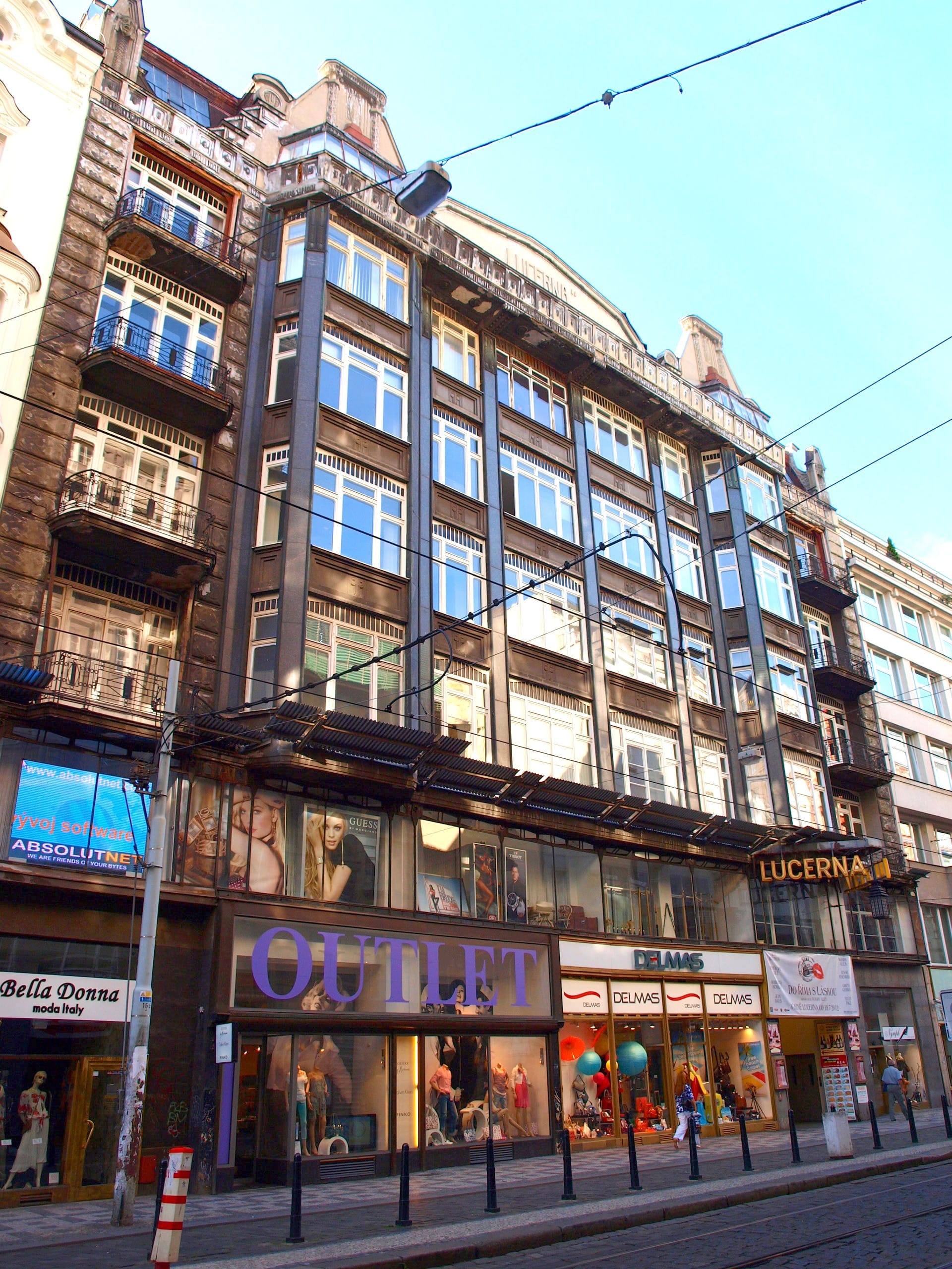 Fassade des Lucerna-Palastes in Prag mit großen Jugendstil-Fenstern, Geschäften im Erdgeschoss und einem klaren blauen Himmel.
