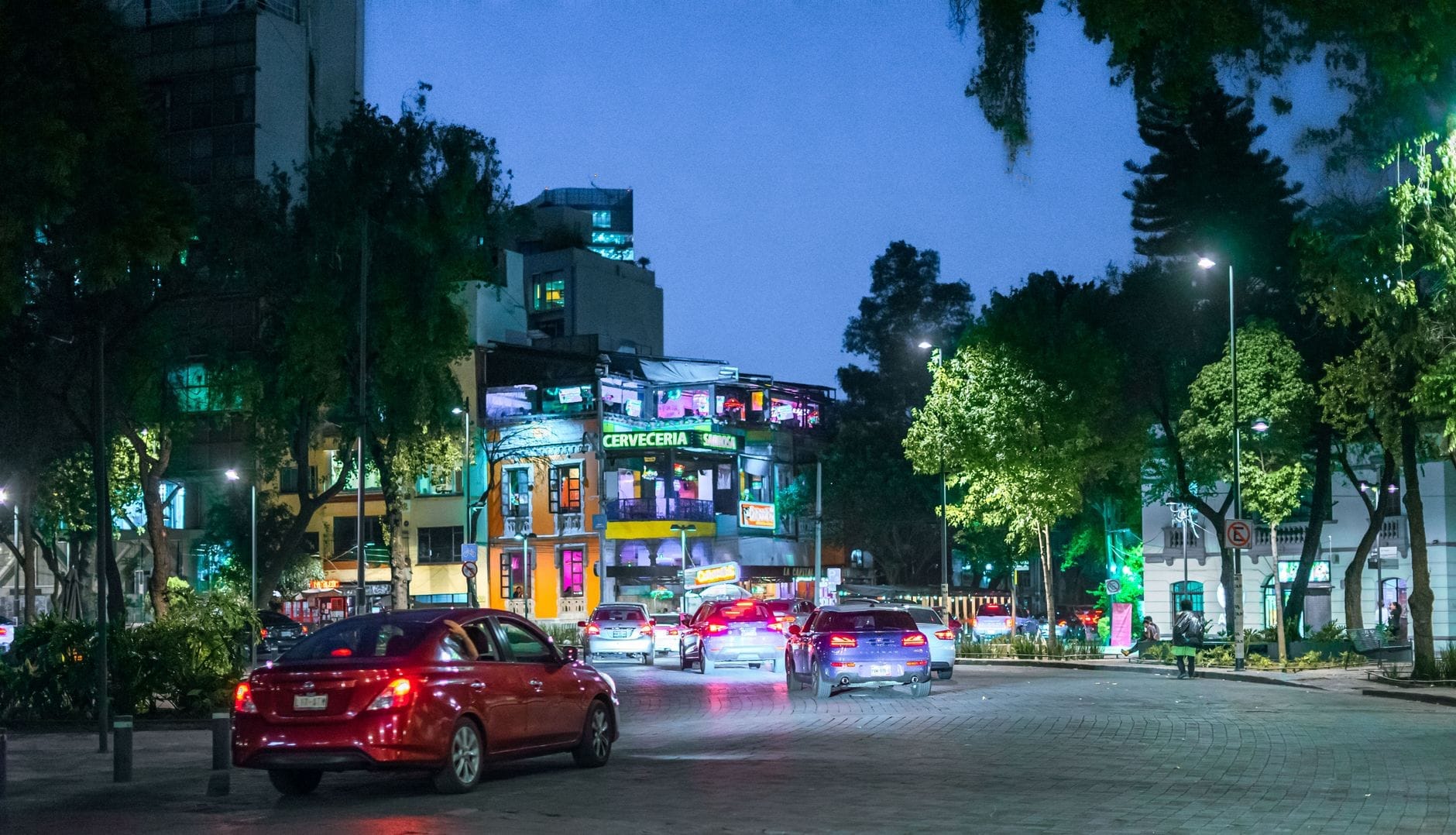 Vista de rua de um animado bairro de Mexico City à noite com carros e edifícios iluminados, sugerindo a vida noturna da cidade.