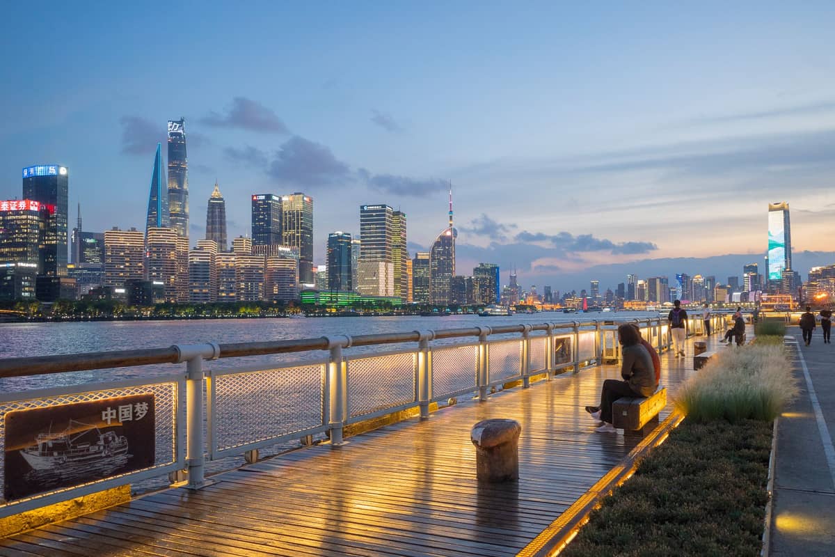 Abendansicht von der Lujiazui Uferpromenade mit dem beleuchteten Holzsteg entlang des Huangpu-Flusses und der Bund-Skyline Shanghais im Hintergrund.