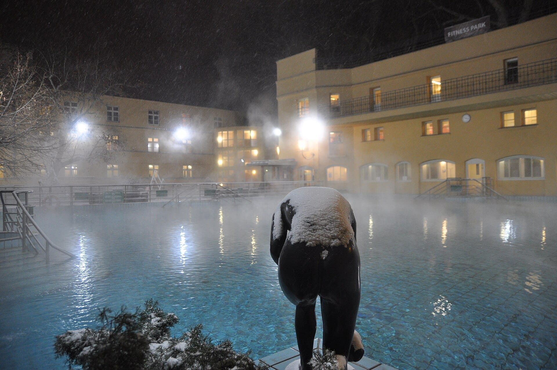 Schneebedeckte Statue mit Blick auf das dampfende Außenbecken im Lukács Thermal Bath, mit warm leuchtenden gelben Gebäuden in der Nacht.