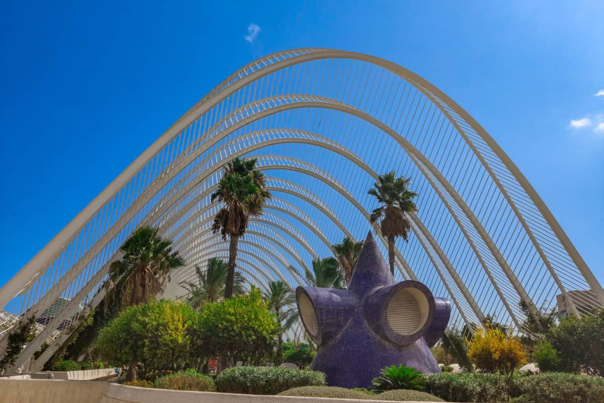 Struttura a pergola con archi bianchi, palme, piante rigogliose e una distintiva scultura viola all'interno di L'Umbracle, nella Città delle Arti e delle Scienze di Valencia, in una giornata di sole.
