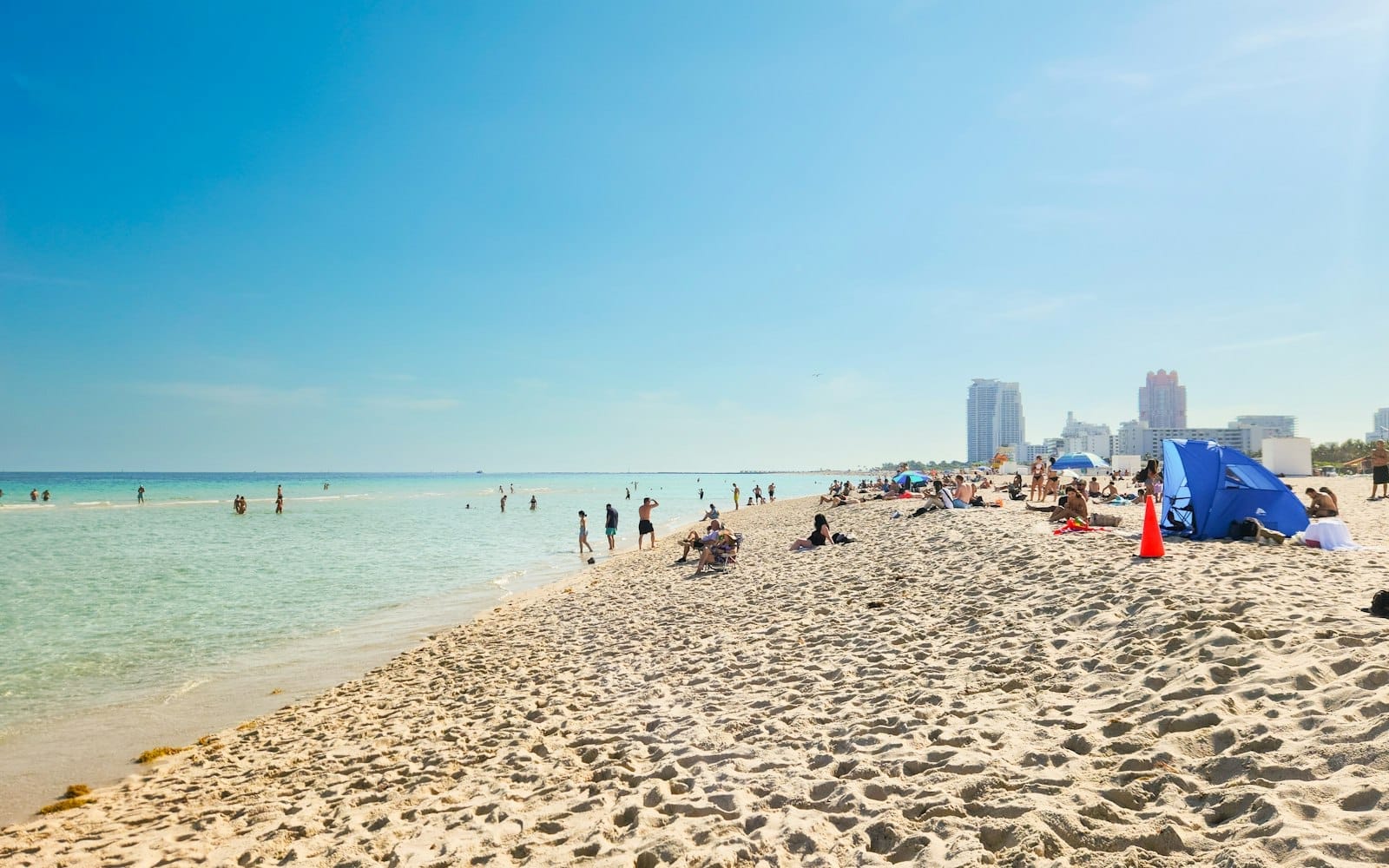 Una vista panoramica di Lummus Park Beach con cielo azzurro, acqua turchese, sabbia chiara e persone che si rilassano e nuotano con lo skyline di Miami Beach sullo sfondo.