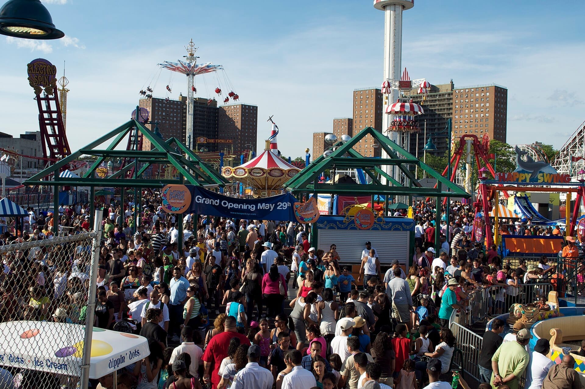 Grandi folle riempiono il Luna Park a Coney Island in una giornata di sole, circondate da giostre colorate, striscioni ed edifici cittadini sullo sfondo.