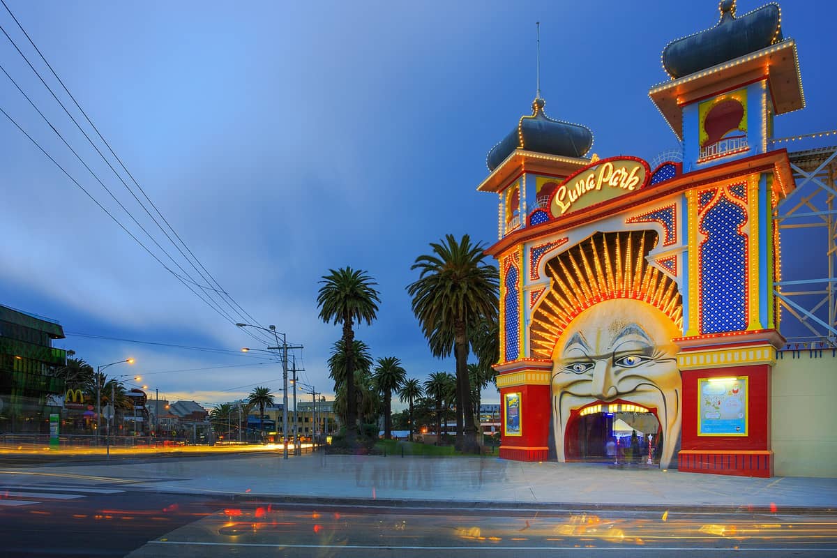 Entrada de Luna Park Melbourne al atardecer, con la famosa fachada iluminada de la boca sonriente y luces vibrantes sobre un cielo azul de noche.