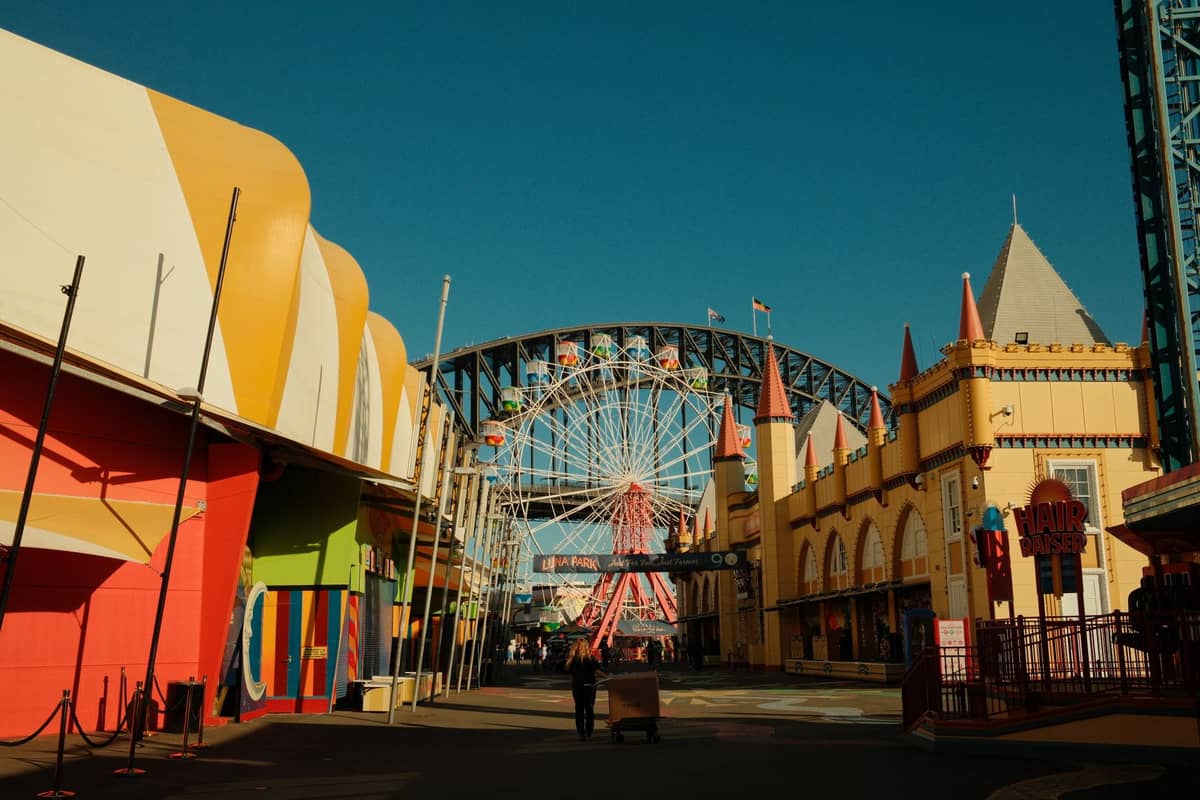 Główna aleja wewnątrz Luna Park Sydney z kolorowymi zabytkowymi budynkami, diabelskim młynem i Sydney Harbour Bridge wyginającym się nad nimi w tle w pogodny dzień.