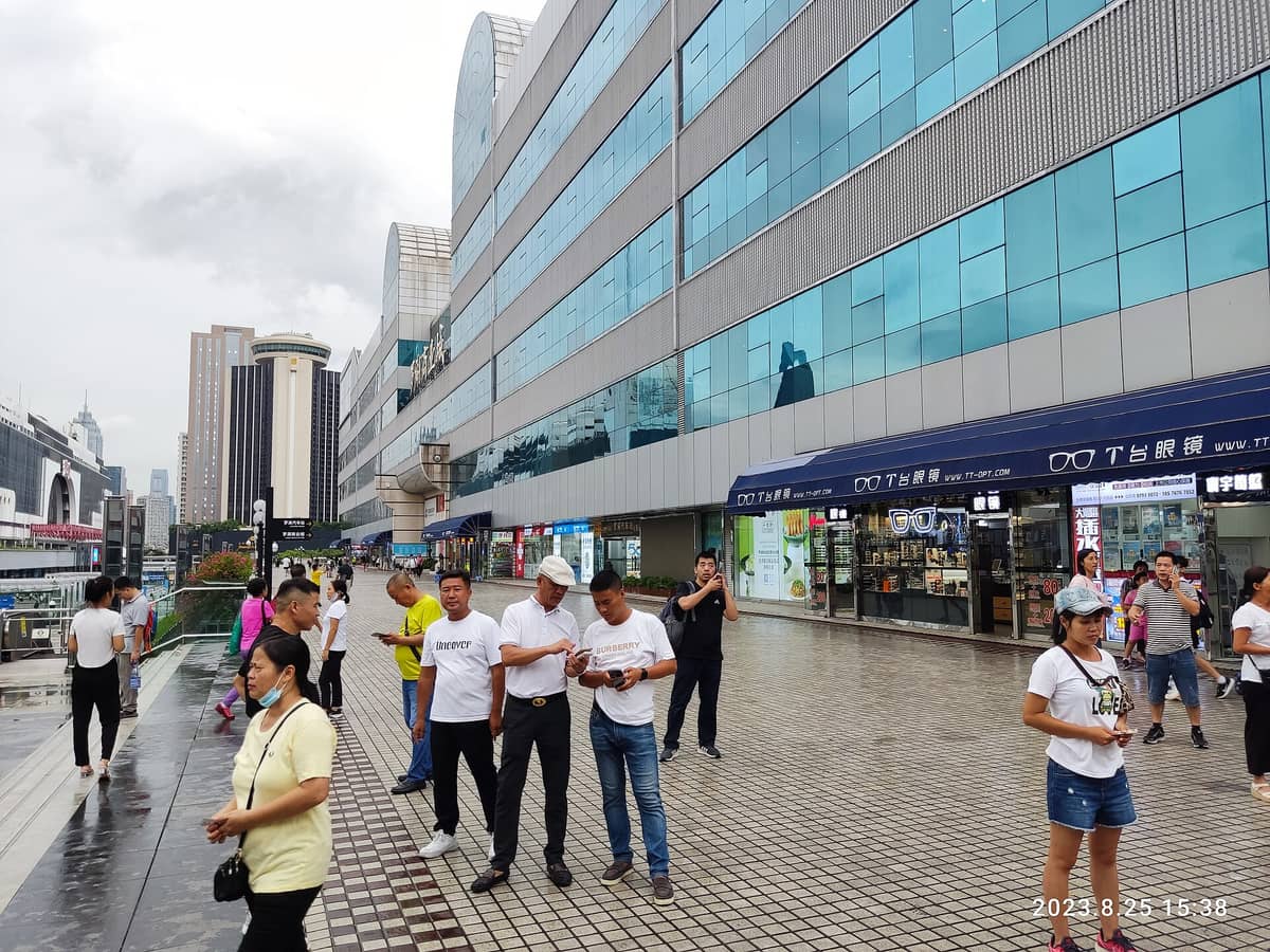 Vista externa do complexo comercial Luohu Commercial City em Shenzhen, com pessoas caminhando na frente e diversas lojas no nível da rua ao longo do edifício.