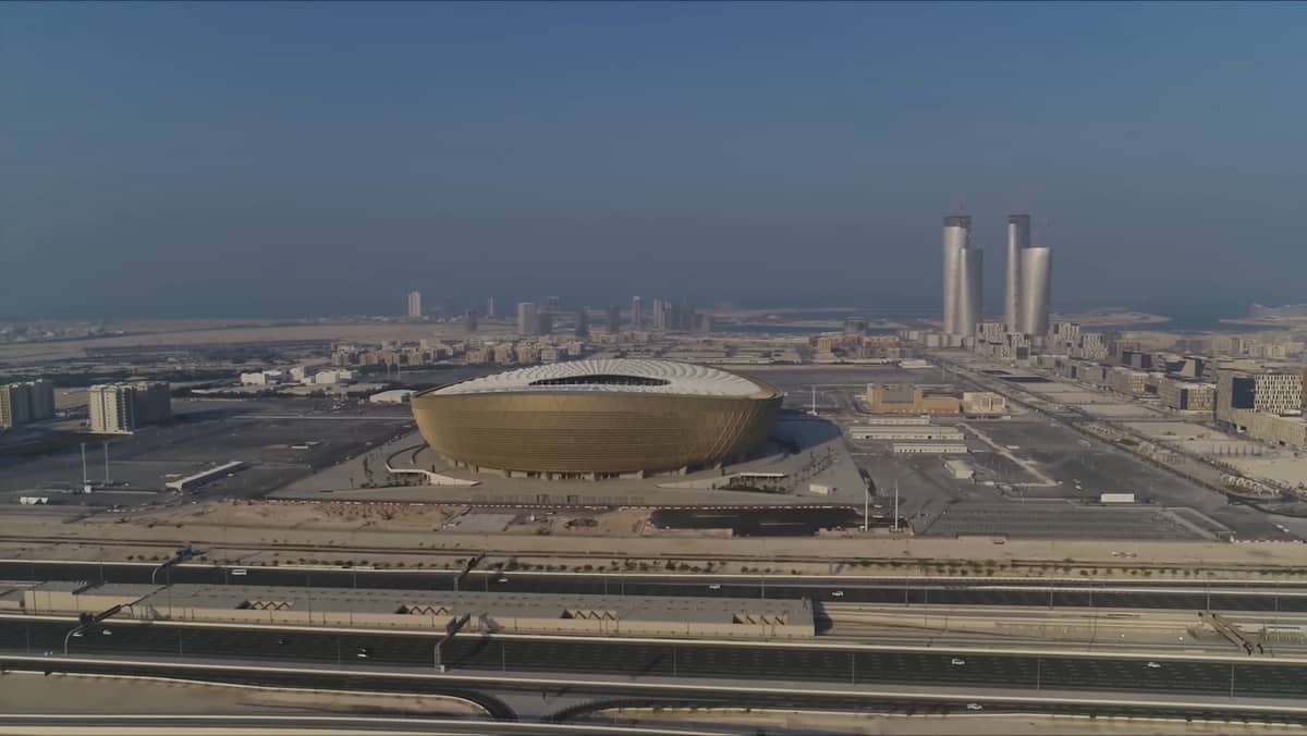 ภาพถ่ายทางอากาศของ Lusail Iconic Stadium ในกาตาร์ โดดเด่นด้วยภายนอกสีทองและสีขาว ล้อมรอบด้วยทิวทัศน์เมืองสมัยใหม่และอาคารสูง