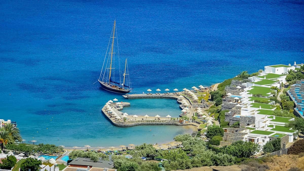 Vista aerea di un resort di lusso sul mare a Creta con piscine private, aree relax e una barca a vela ancorata nel mare blu cristallino.