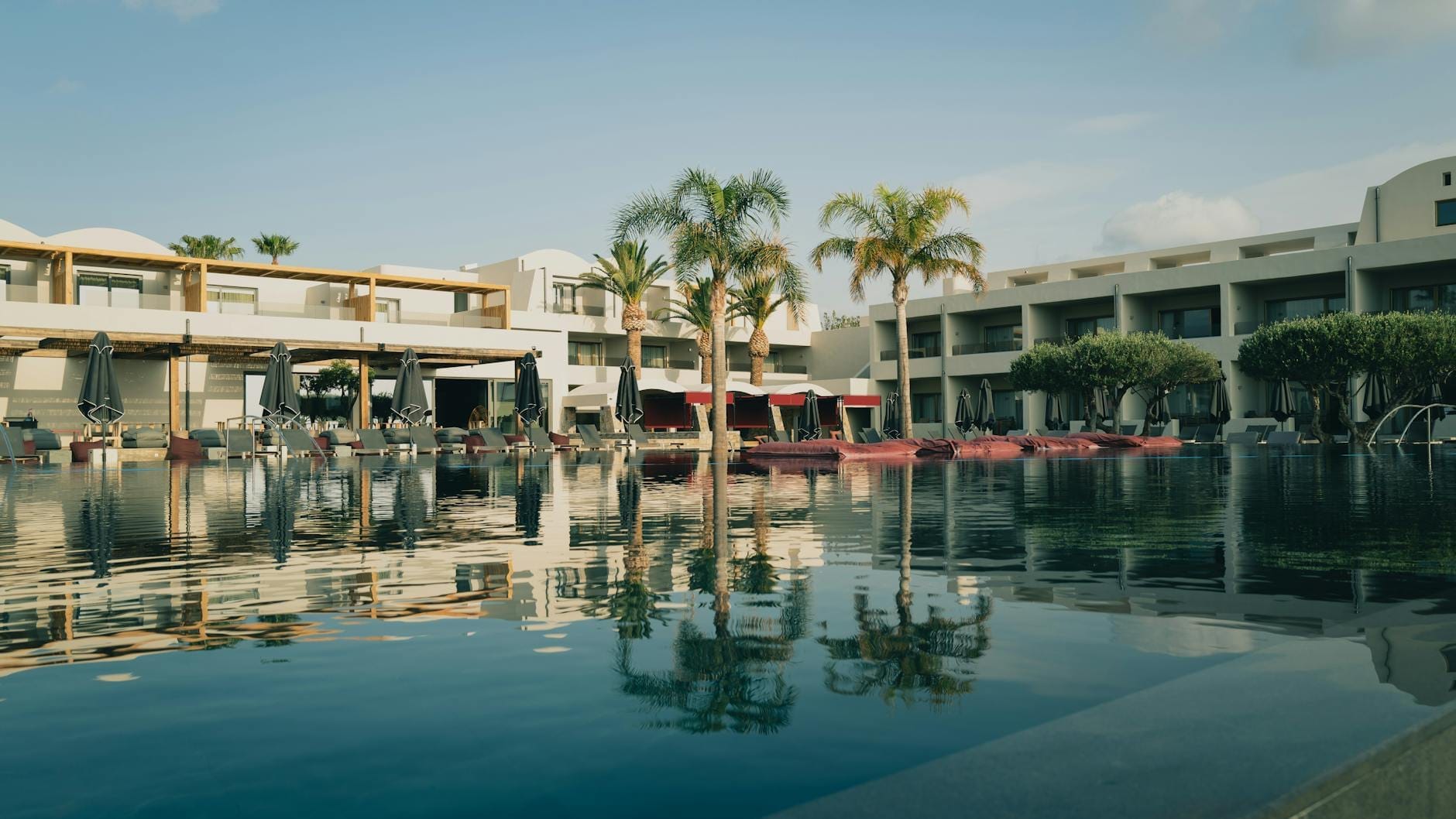 Resort de luxo com arquitetura moderna, palmeiras e espreguiçadeiras refletidas em uma piscina serena sob o céu azul de Creta.