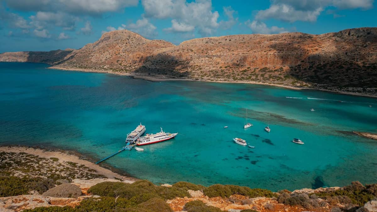 Baia turchese a Creta con yacht di lusso e una grande imbarcazione ormeggiata vicino a una costa rocciosa sotto un cielo azzurro.