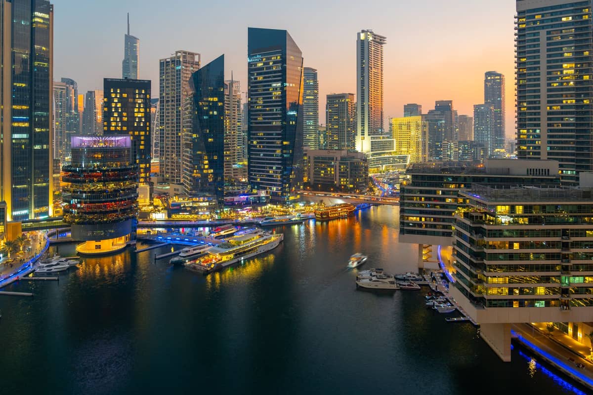 Luftaufnahme der Dubai Marina bei Sonnenuntergang mit Luxusyachten, Hochhäusern, gleißenden Lichtern und lebhafter Uferpromenade.