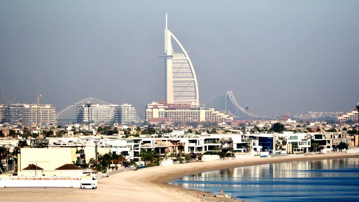 Das ikonische Burj Al Arab Hotel in Dubai ragt markant vor der Stadtsilhouette auf, mit Strandvillen und ruhigem blauen Wasser im Vordergrund.