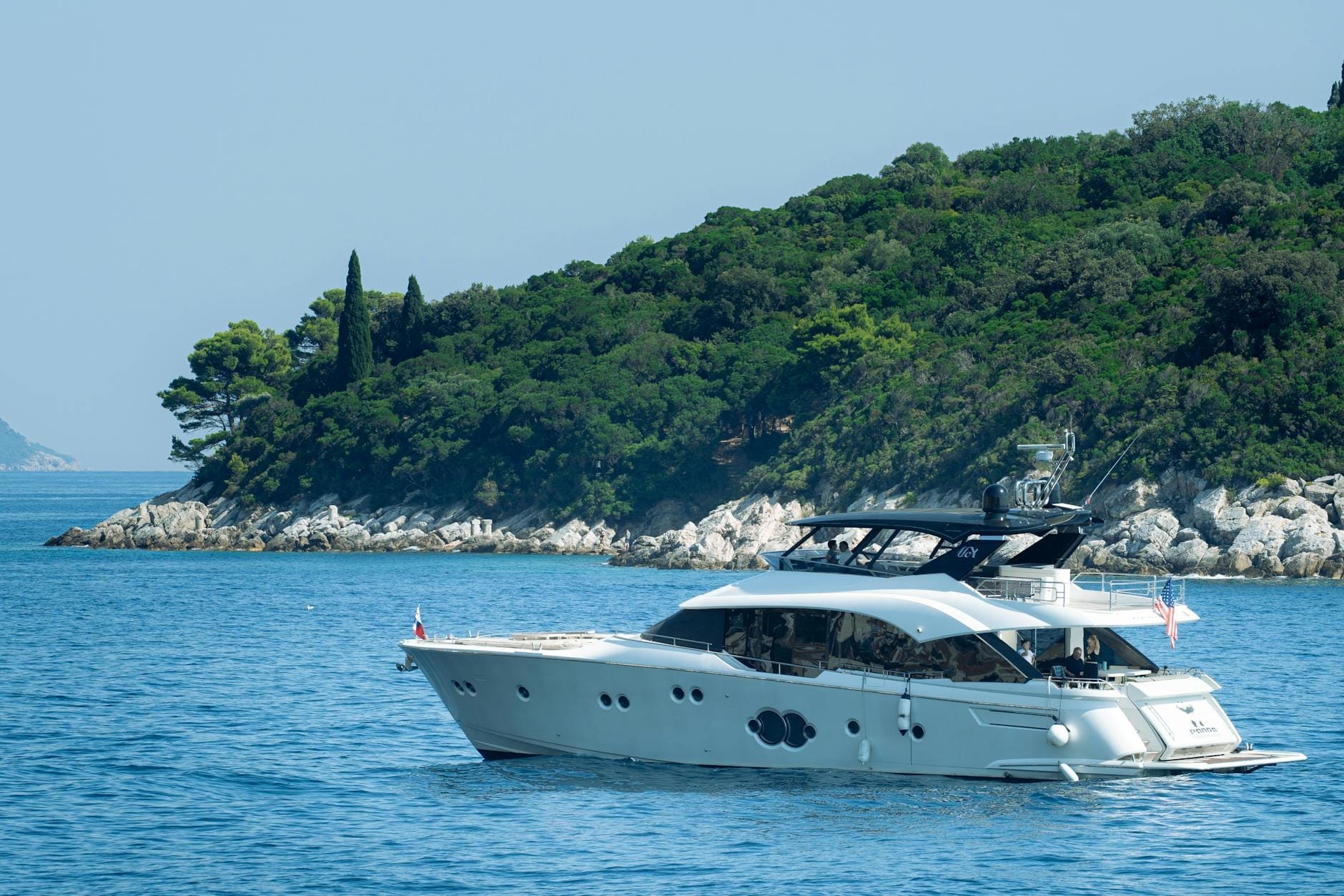 Yate de lujo navegando por aguas azules en calma con una exuberante isla verde al fondo, ilustrando una experiencia náutica de lujo en Dubrovnik.