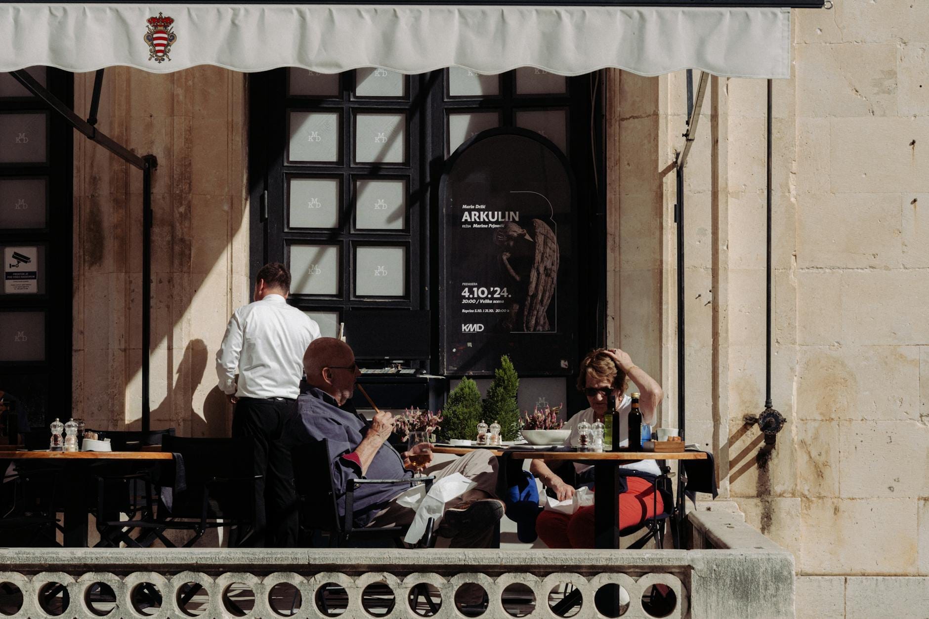 Escena de comedor al aire libre en un restaurante elegante con un camarero atendiendo a los clientes, frente a la histórica arquitectura de piedra de Dubrovnik.