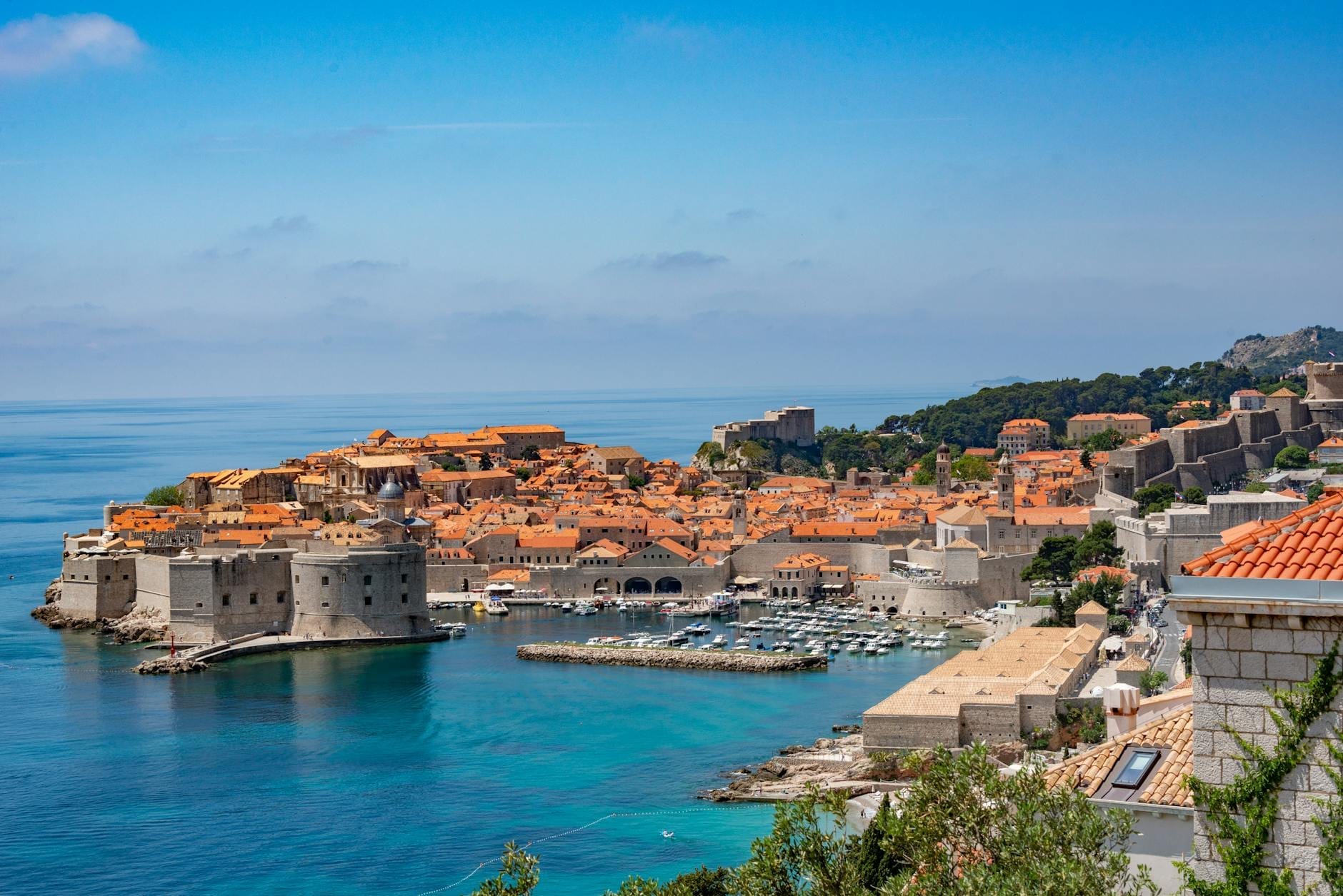 Vista panorámica de las murallas de piedra y el puerto del casco antiguo de Dubrovnik, con el mar azul claro y edificios históricos bajo un cielo luminoso.