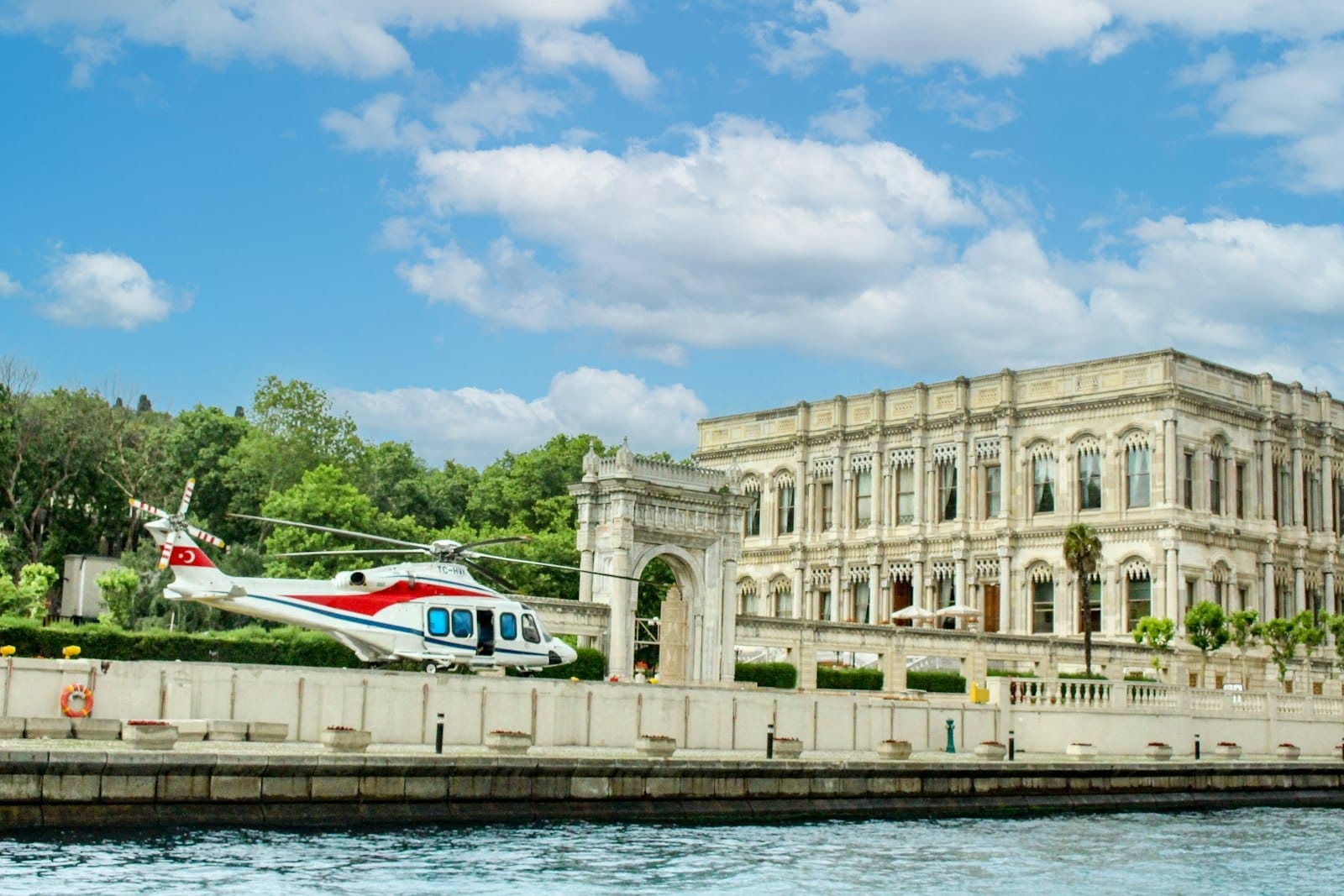 Palácio neoclássico elegante à beira do Bósforo, com jardins exuberantes e um helicóptero particular no heliporto, sob um céu azul brilhante com nuvens fofas.