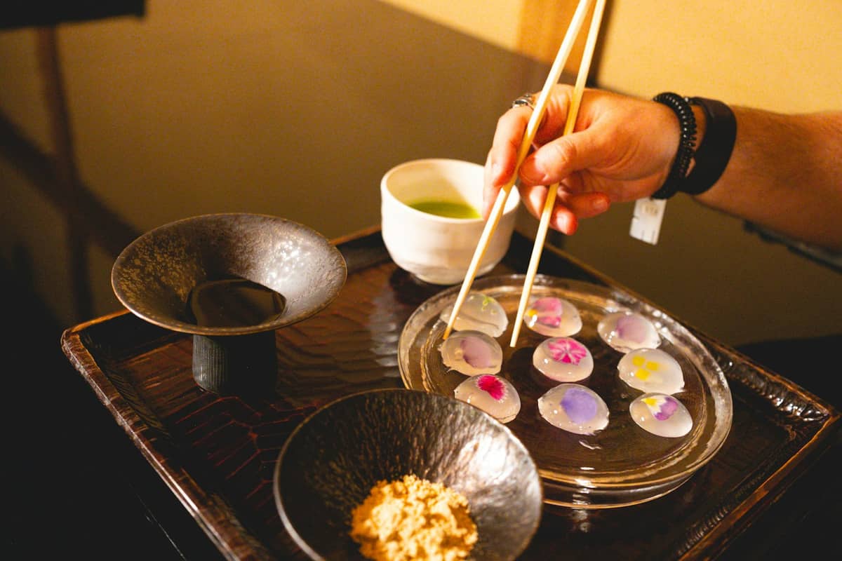 A hand with chopsticks selects a delicate plate with artfully arranged bite-sized dishes and a cup of green tea on a wooden tray.