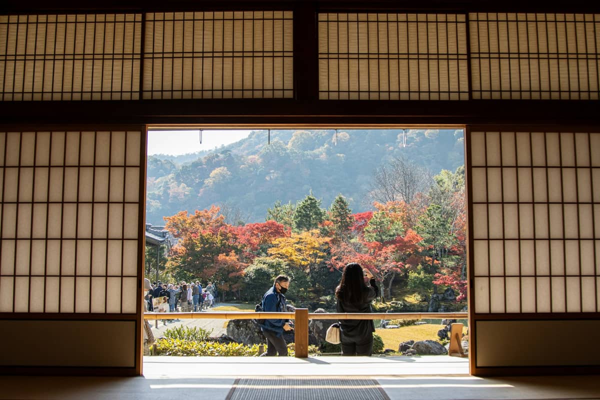 Looking out from a traditional tatami room in a Kyoto ryokan at autumn trees and a tranquil Japanese garden with people enjoying the view.