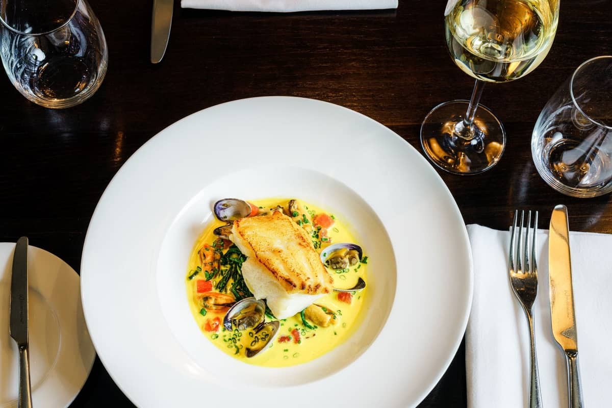 Prato elegante de peixe com amêijoas e uma taça de vinho branco numa mesa de restaurante de alta gastronomia.
