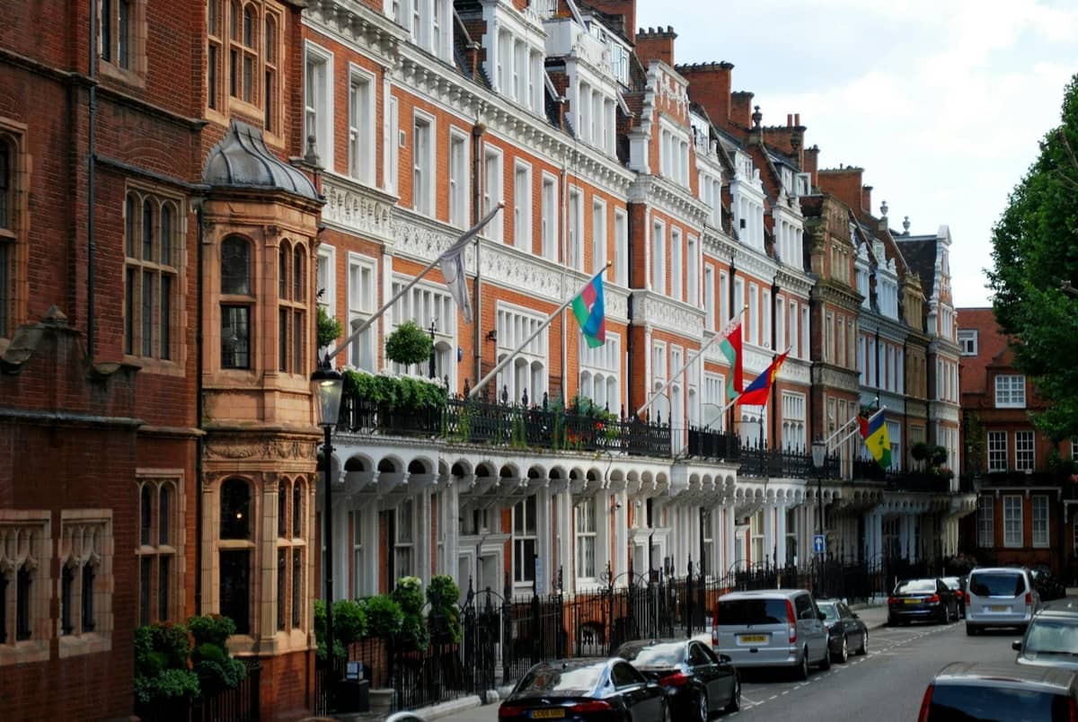 Elegantes casas em terraço com bandeiras, varandas ornamentadas e carros de luxo numa rua tranquila de um bairro sofisticado de Londres.