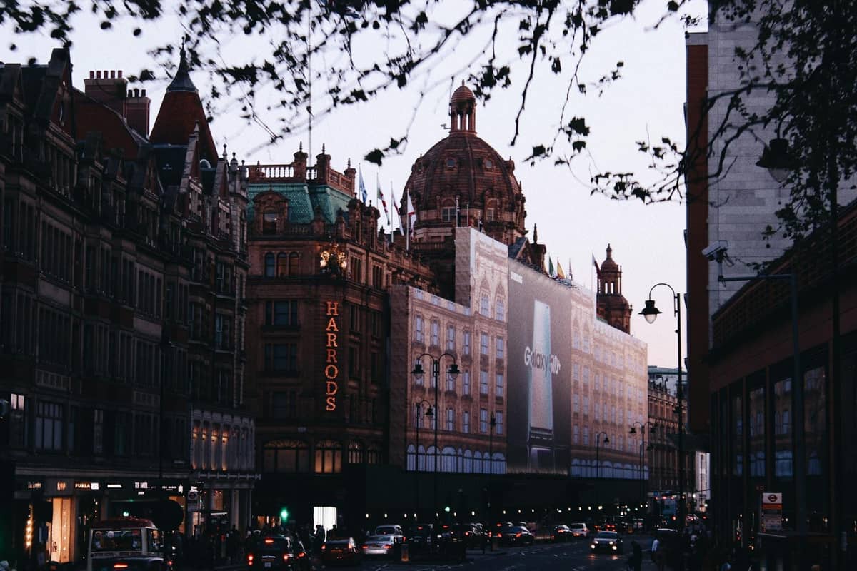 Vista da rua dos grandes armazéns de luxo Harrods em Londres ao entardecer, com arquitetura clássica e tráfego intenso na frente.