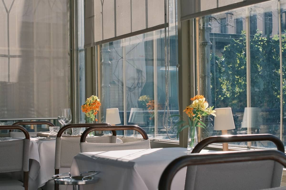 Interior elegante de restaurante con manteles blancos, flores frescas, cristalería y grandes ventanales que dejan entrar la luz del día, sugiriendo un ambiente de comedor de lujo.