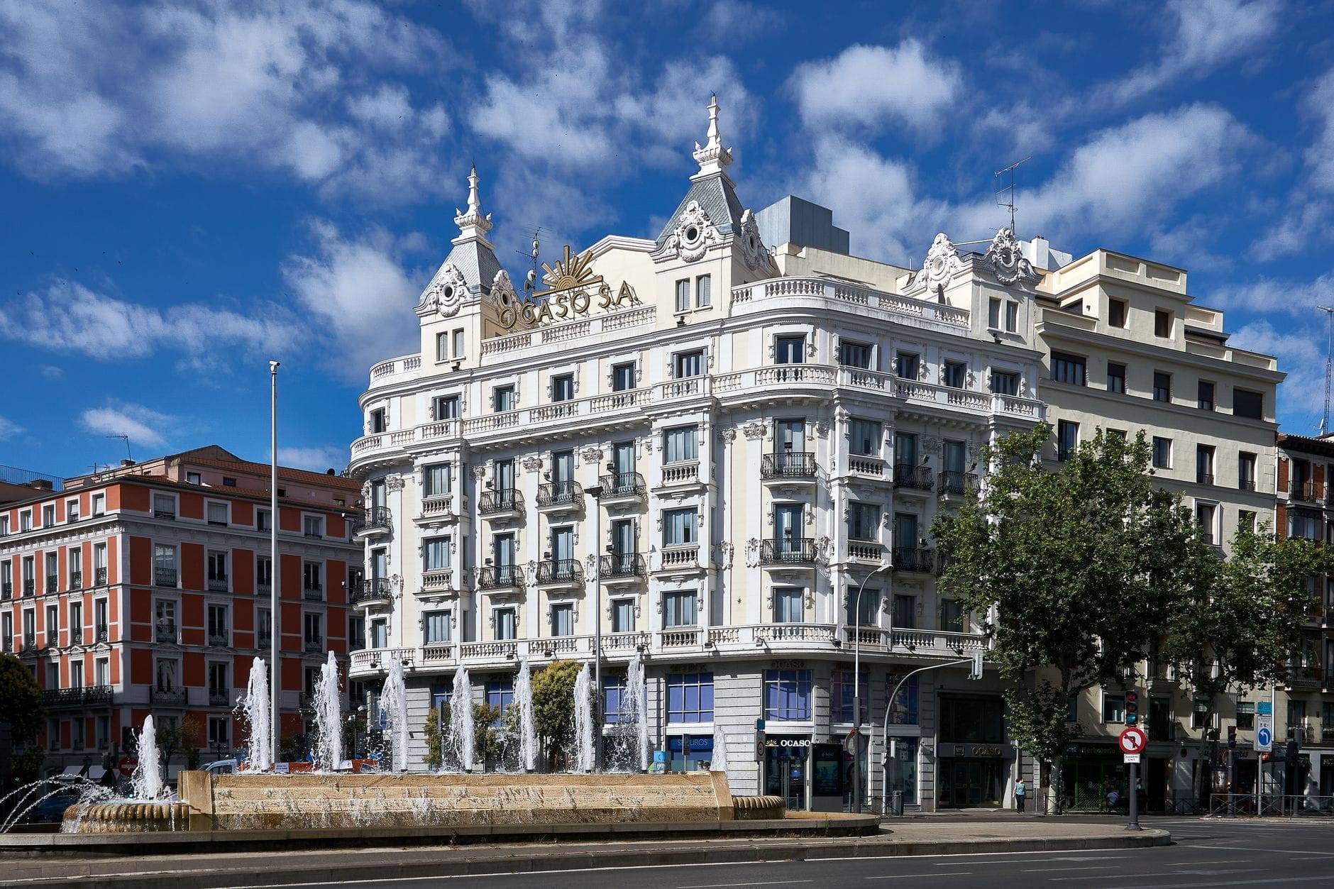 Elegante edifício histórico branco em Madri com sacadas ornamentadas e fachada decorada, visto de uma praça com fontes e céu azul ao fundo.