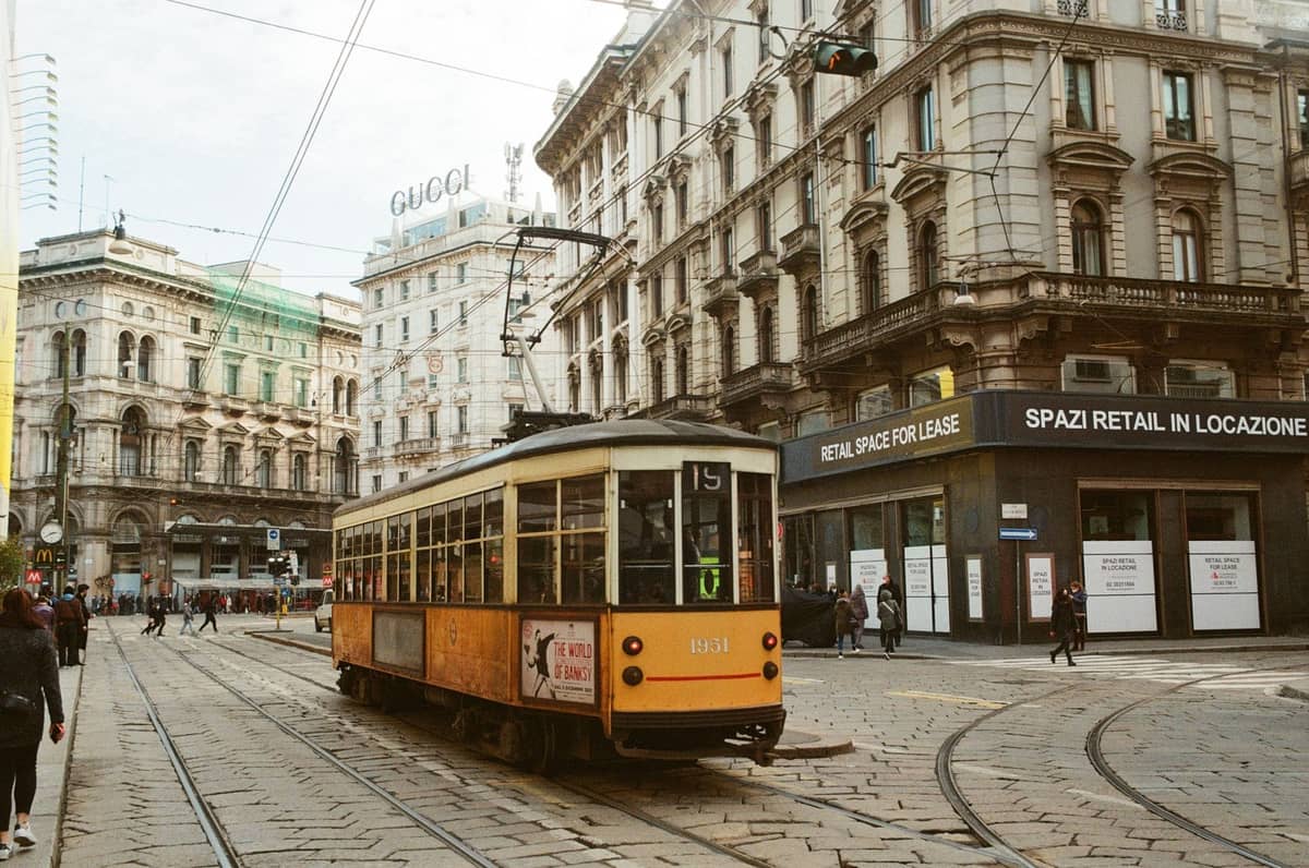 Klasyczny żółty tramwaj jedzie przez luksusową dzielnicę Mediolanu z eleganckimi historycznymi budynkami, szyldem GUCCI i ekskluzywnymi sklepami.