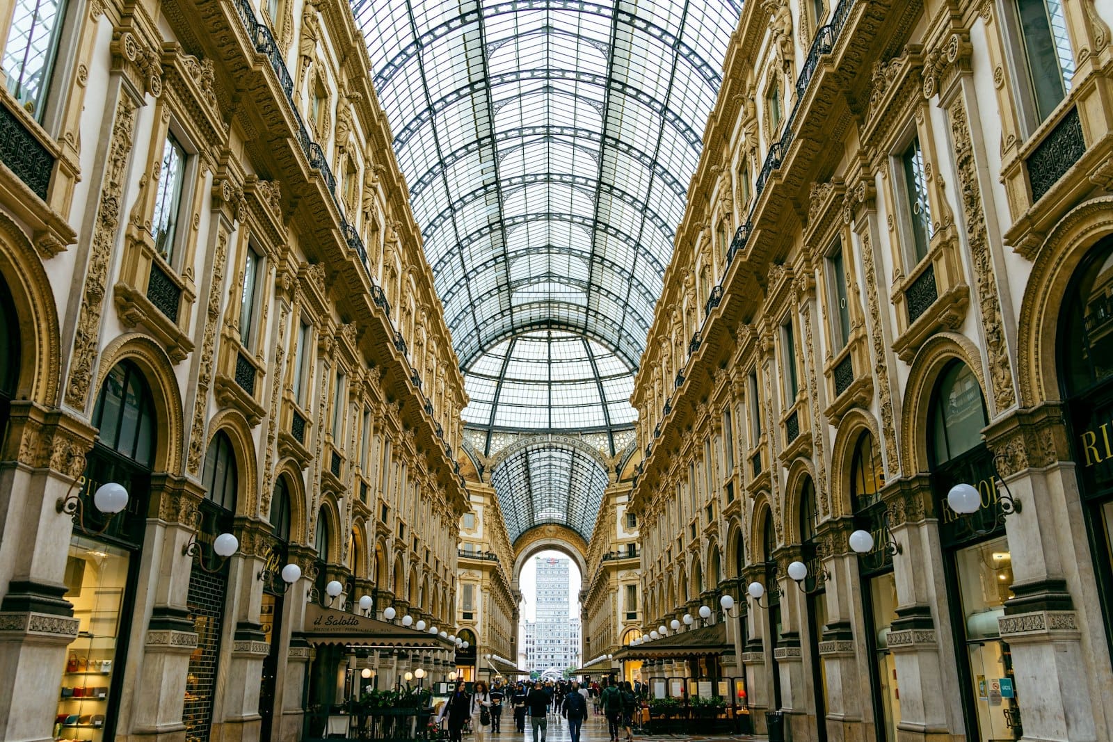 Vista ampla da luxuosa Galleria Vittorio Emanuele II em Milão, com seus arcos ornamentados, lojas elegantes e um imenso teto de vidro repleto de luz.