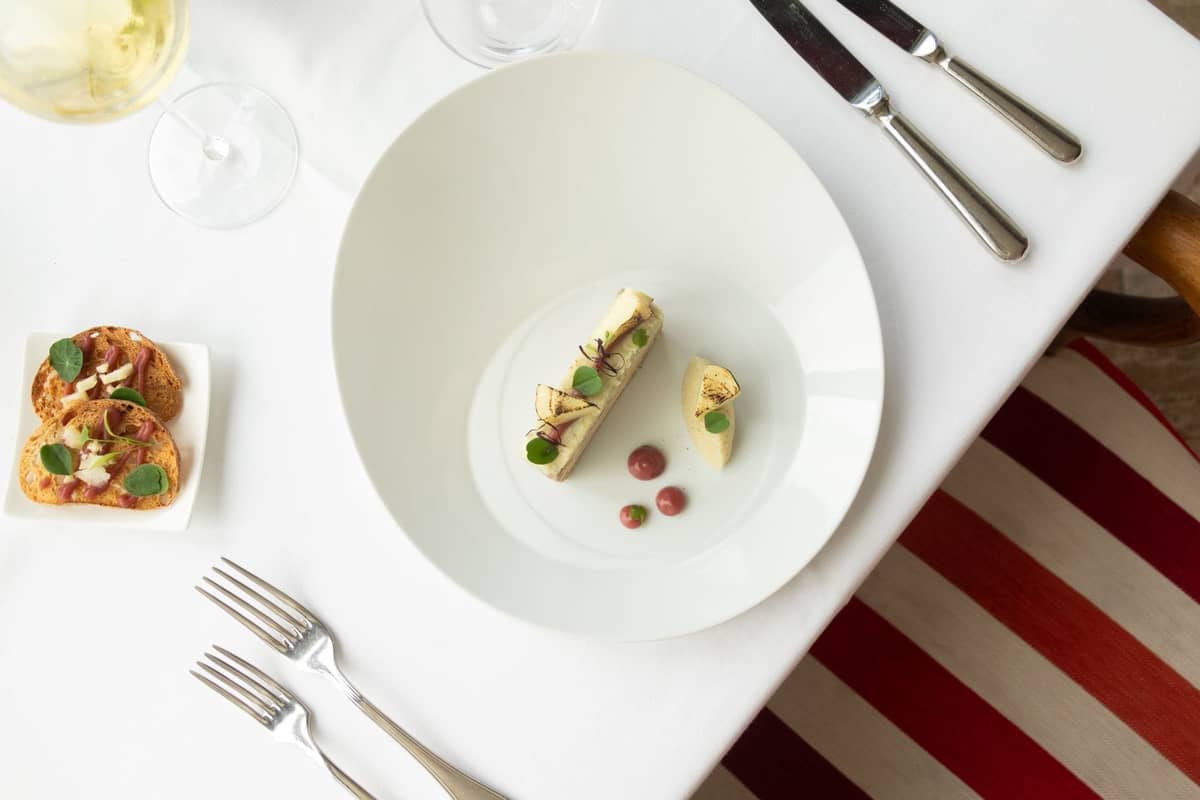 Elegantes angerichtetes Gericht und Amuse-bouches auf einem weißen Tischtuch, mit Weinglas und Besteck in einem Gourmetrestaurant.