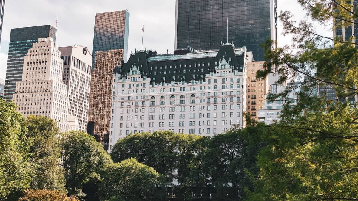 Eleganter Blick auf das Plaza Hotel und die Wolkenkratzer von Midtown, die hinter den belaubten Bäumen des Central Park in Manhattan, New York City, aufragen.