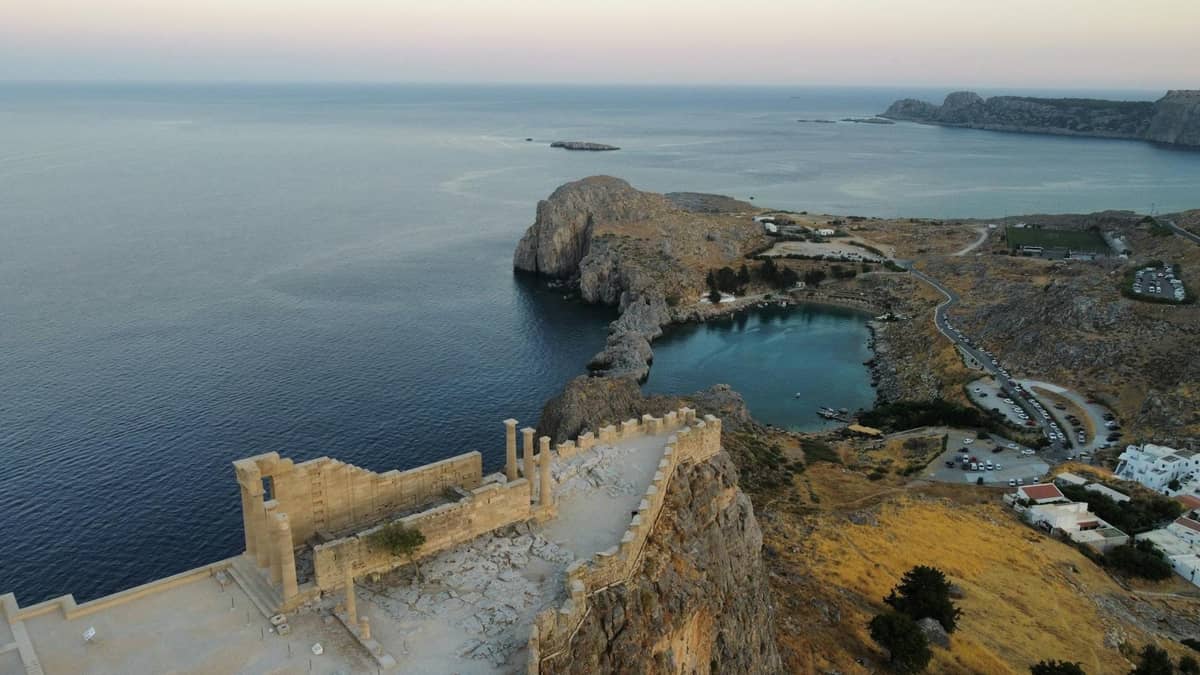 Aerial view of the Acropolis of Lindos on a cliff above the sea, with ancient ruins and a turquoise bay below.