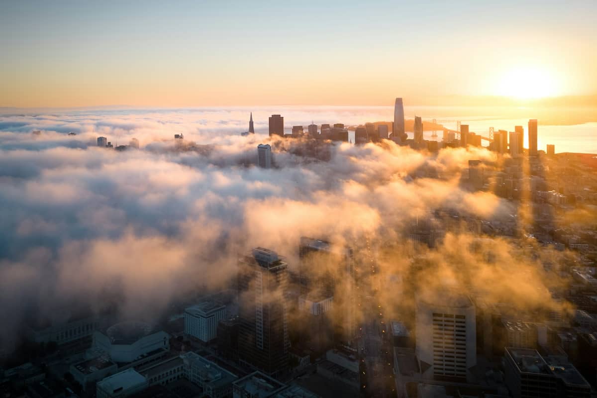 Una spettacolare vista aerea dello skyline del centro di San Francisco con grattacieli iconici che emergono da una nebbia dorata all'alba, rappresentando l'atmosfera urbana compatta e di lusso della città.