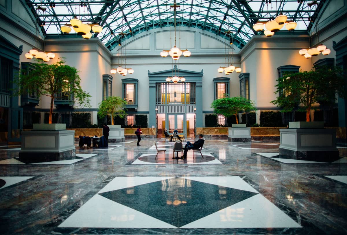 Grande lobby con pavimenti in marmo, soffitti alti, grandi finestre e un tetto in vetro con atrio, con eleganti sedute e alberi in vaso.