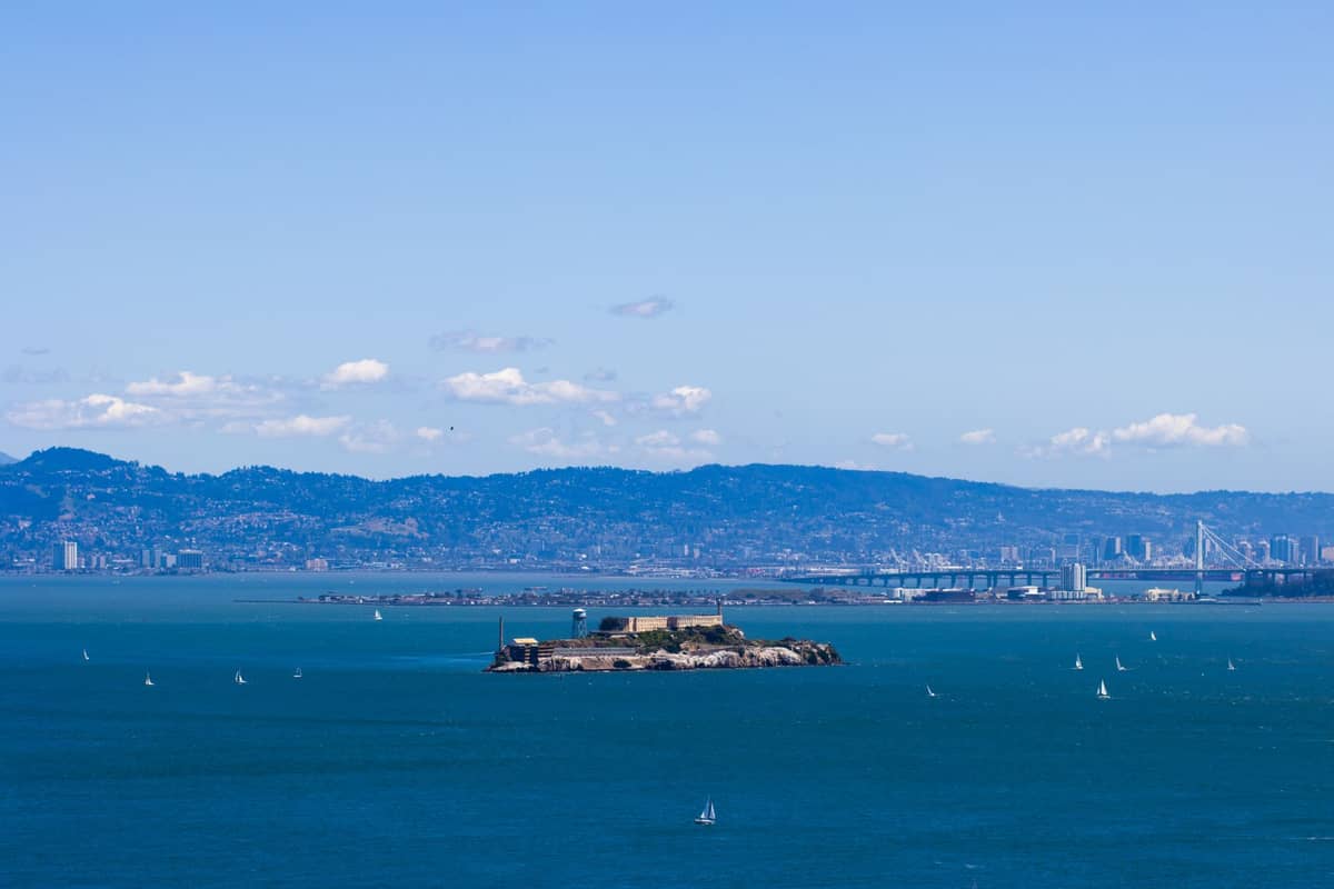 Vista panoramica dell'isola di Alcatraz circondata da acqua blu, con la città di San Francisco e le colline sullo sfondo sotto un cielo azzurro limpido.