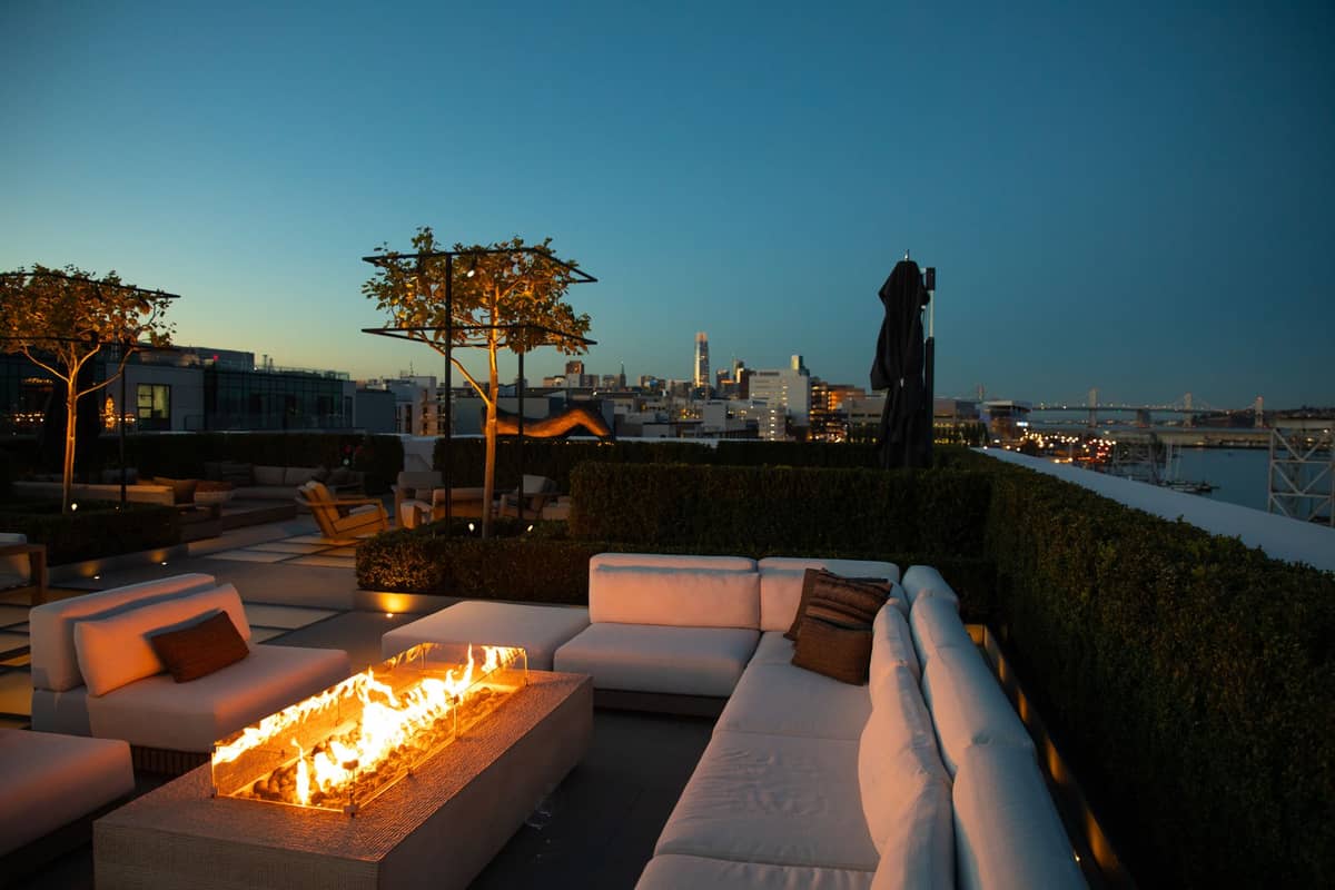 Elegante terraza de lujo al atardecer con chimenea moderna, sillones mullidos y vistas panorámicas del centro de San Francisco al fondo.