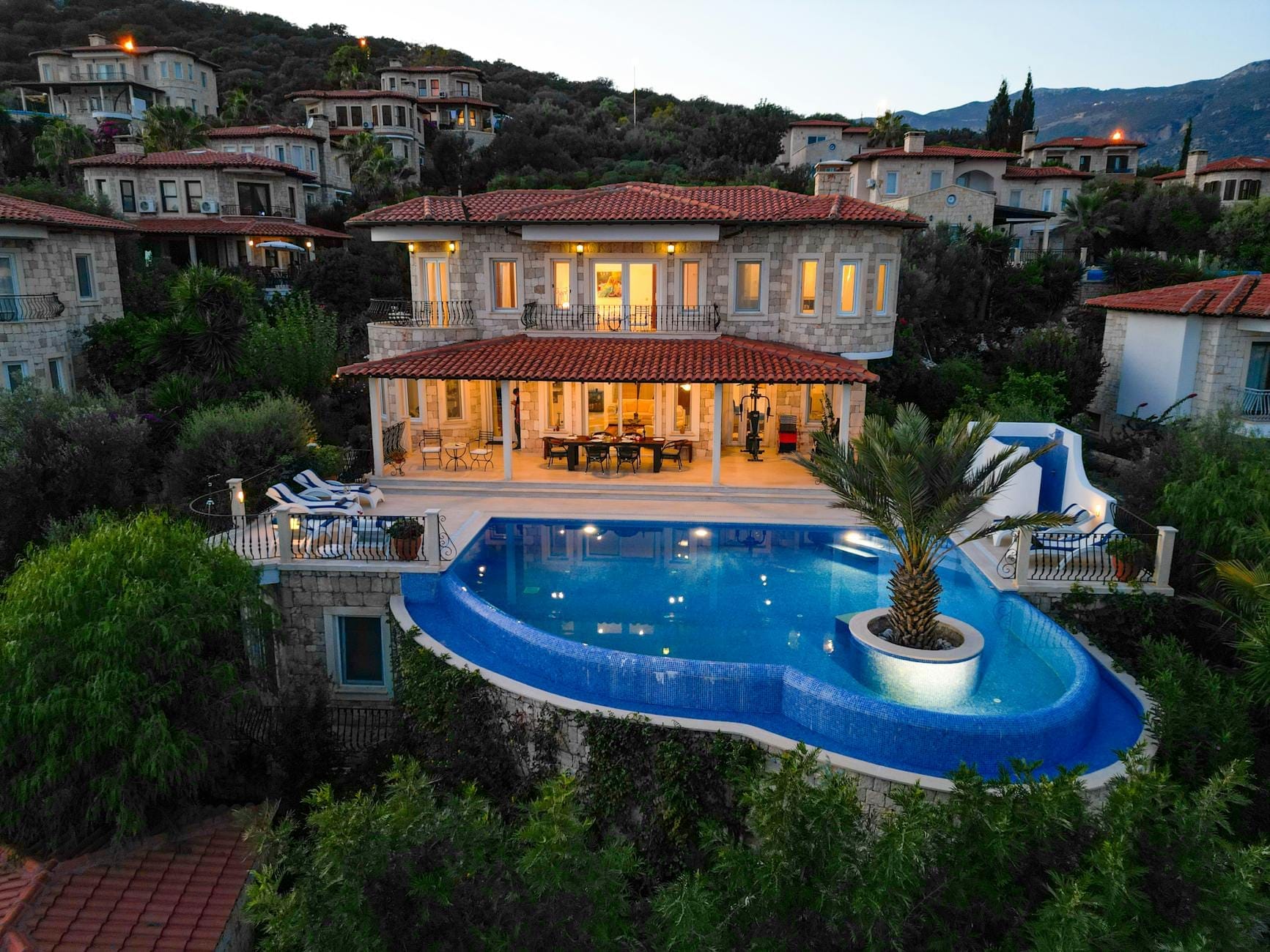 Eine große, luxuriöse Villa mit privatem Infinity-Pool, umgeben von Grün und Hangvillen, bei Sonnenuntergang oder in der Dämmerung.