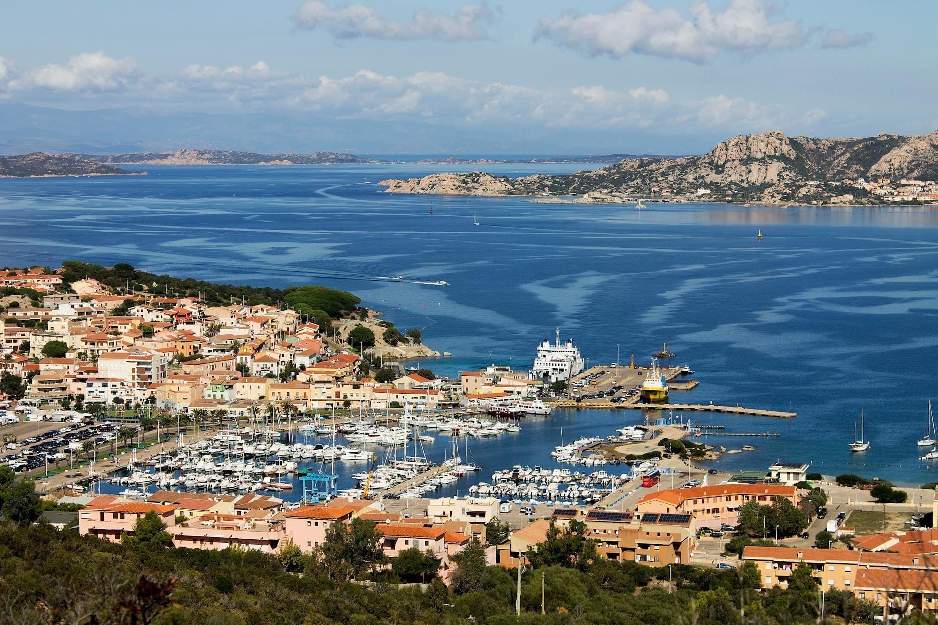 Panoramablick auf einen Yachthafen mit Yachten, Booten und Luxushäusern an der Küste Sardiniens, mit klarem blauem Wasser und felsigen Hügeln.