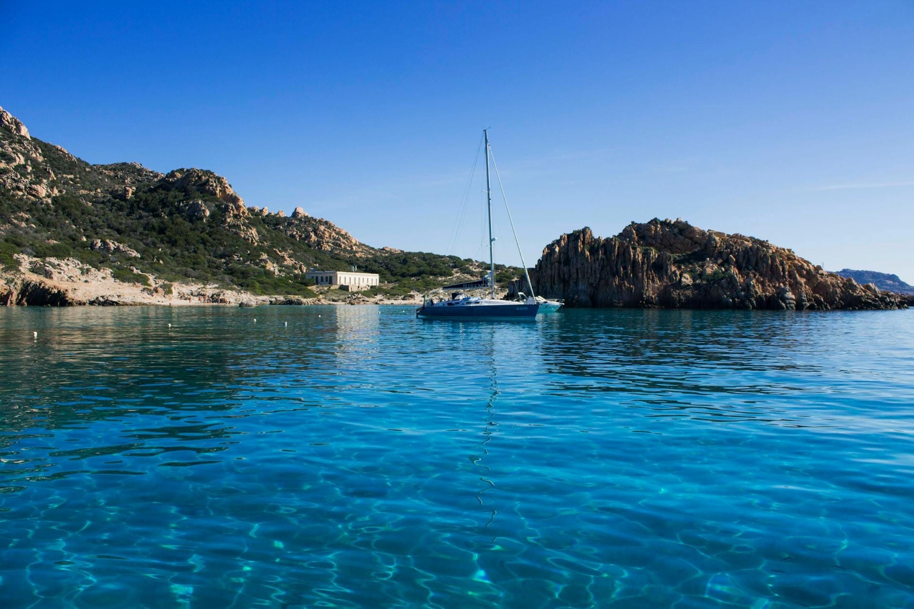 Segelboot in klarem türkisfarbenem Wasser vor der felsigen Küste Sardiniens unter einem strahlend blauen Himmel vor Anker.