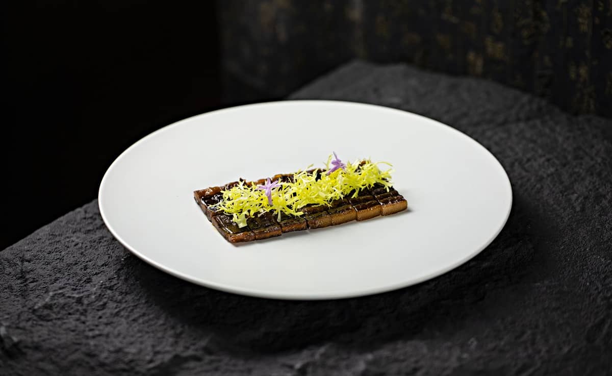 Prato minimalista de alta gastronomia com guarnições elaboradas em um grande prato redondo branco, sobre uma superfície escura semelhante a pedra.