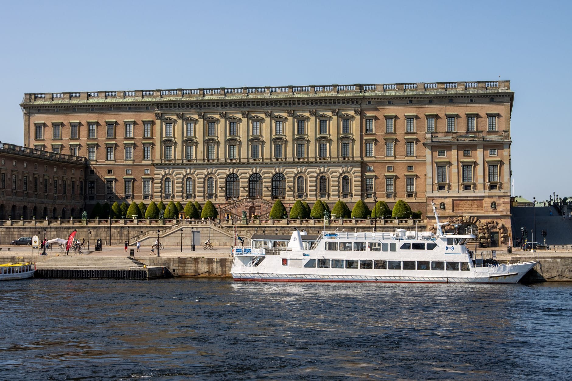พระราชวัง Stockholm Royal Palace ริมน้ำพร้อมเรือนำเที่ยวอยู่ด้านหน้า ภายใต้ท้องฟ้าสีฟ้าแจ่มใส