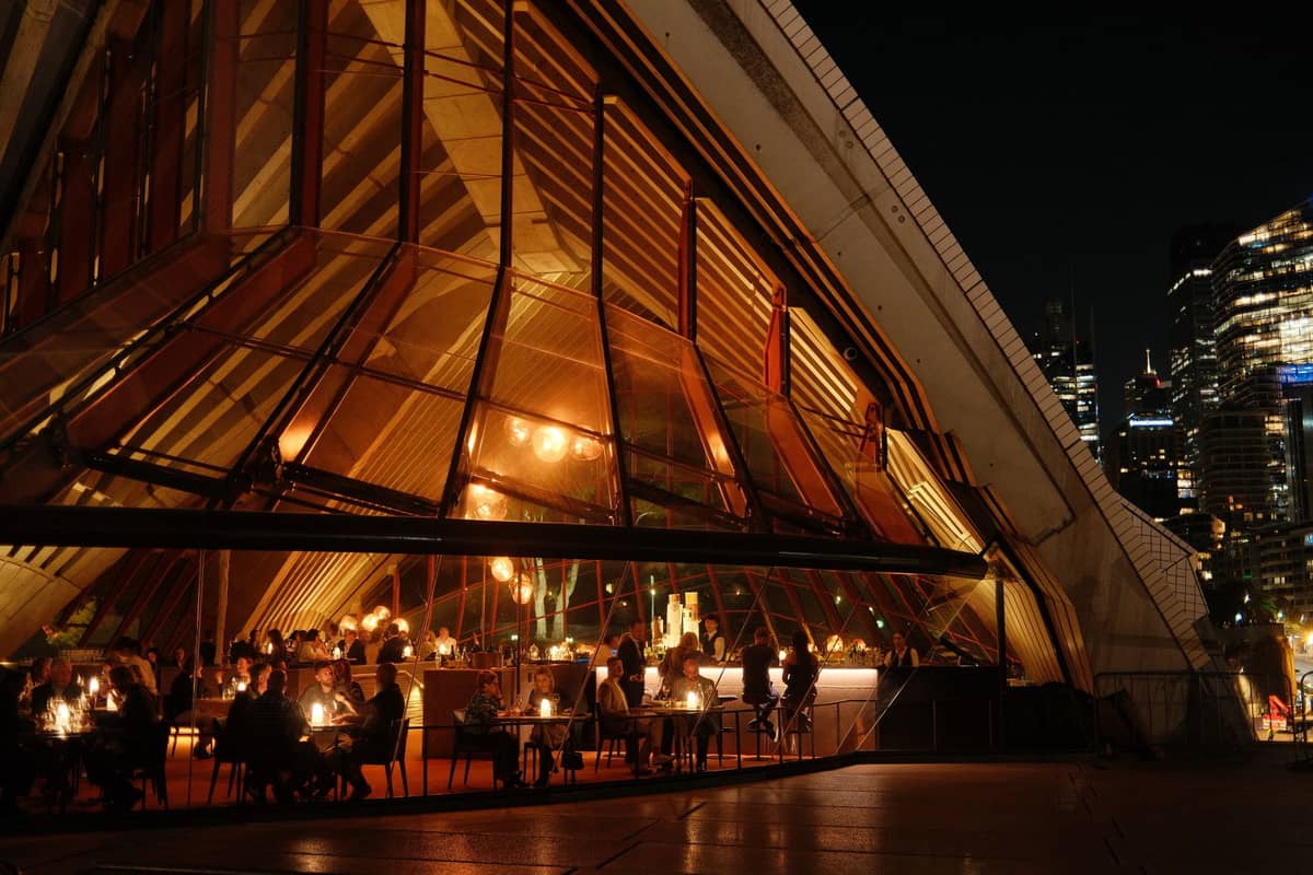 Elegant diners seated at night inside the Sydney Opera House restaurant, with city lights glimmering through angular glass walls.