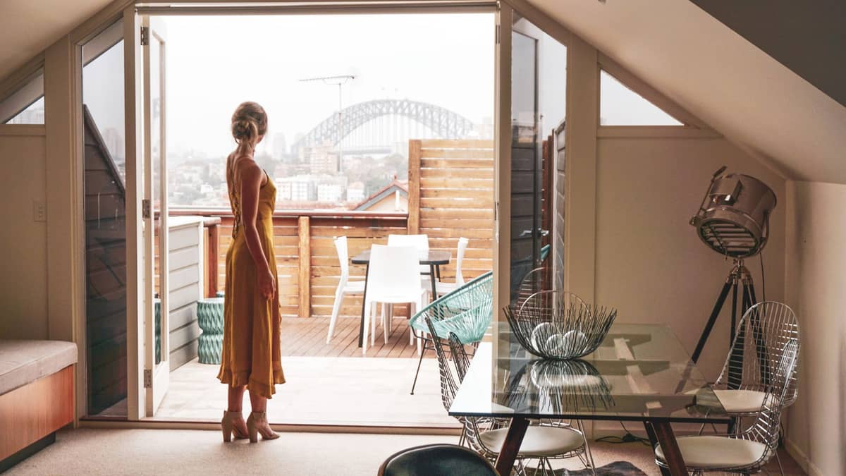 Woman in a luxury Sydney hotel suite with a private terrace, overlooking Sydney Harbour Bridge through open doors, blending high-end accommodation with iconic city views.