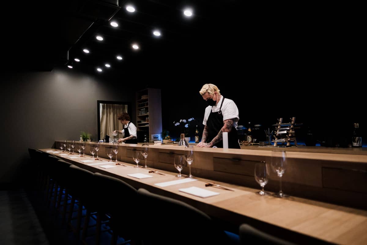 Long wooden chef’s counter set for fine dining, with chefs working in a sophisticated, dimly lit restaurant.