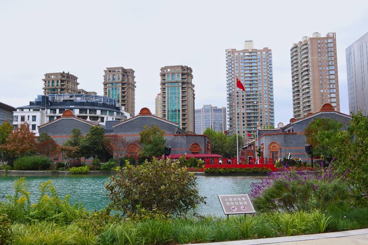 Blick auf einen Teich mit üppigem Grün und historischen Gebäuden im Shanghai-Stil, Hochhäuser im Hintergrund, unter einem hellen Himmel.