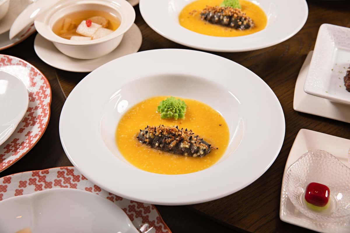 Mehrere elegant angerichtete Gerichte, darunter Seegurke auf gelbem Sauce und feines chinesisches Geschirr, die auf ein gehobenes zeitgenössisches chinesisches Dining hinweisen.