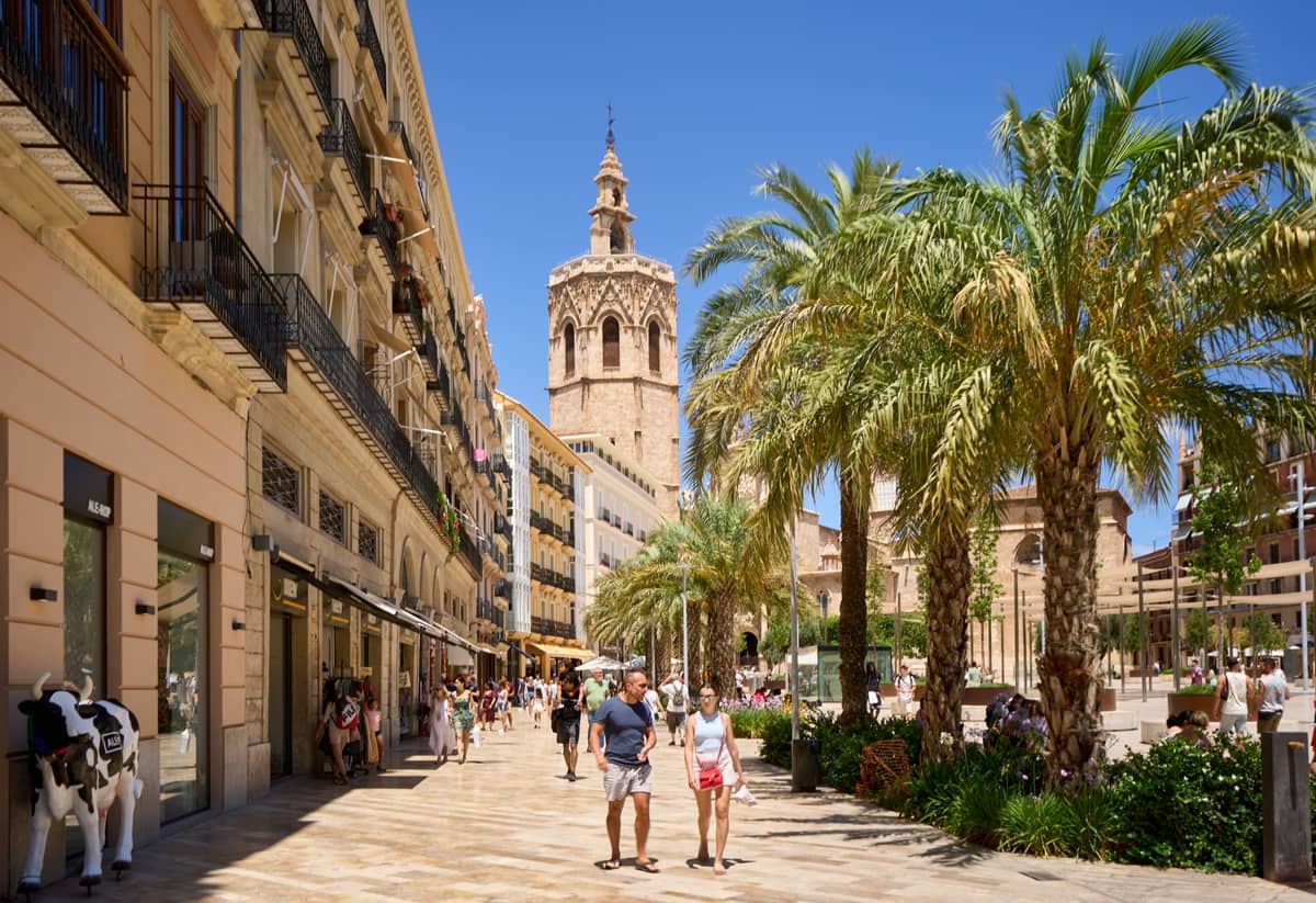 Valencia'nın tarihi merkezinde palmiye ağaçları, yürüyen insanlar ve arka planda ikonik Miguelete kulesi ile geniş yaya caddesi.